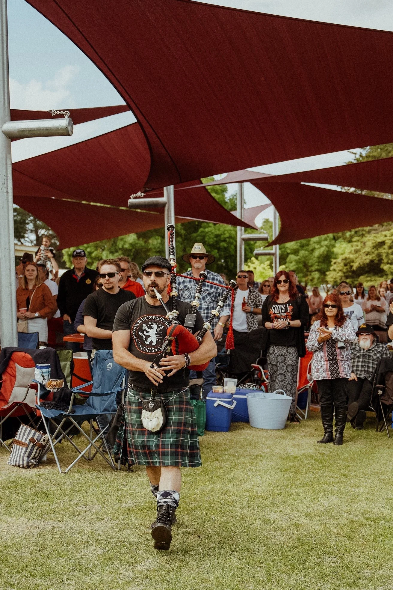 A man in festival attire playing bagpipes at an outdoor event, with a crowd of onlookers under red shade sails.