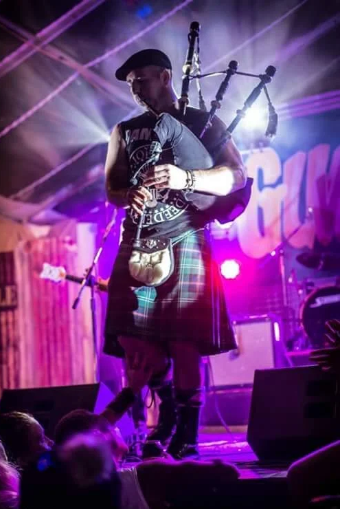 A man dressed in a kilt playing bagpipes on stage with colorful lighting.