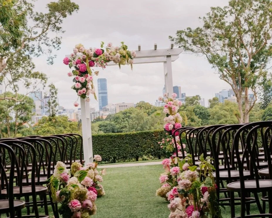 Outdoor wedding ceremony setup with black chairs arranged on either side of a grass aisle leading to a white floral arch with pink and white flowers, overlooking a city skyline with tall buildings and greenery.