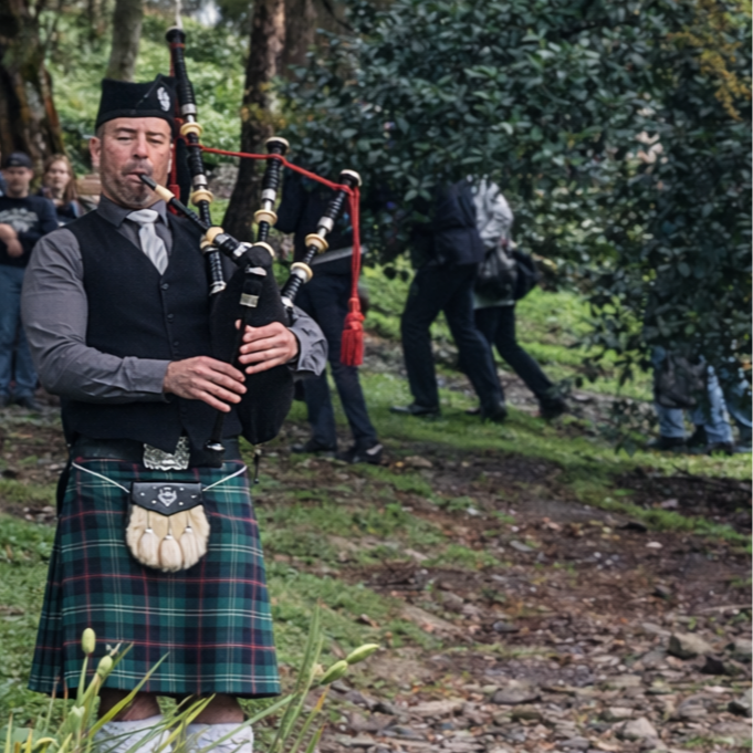 A man wearing traditional Scottish attire, including a plaid kilt, playing the bagpipes outdoors with people walking in the background.