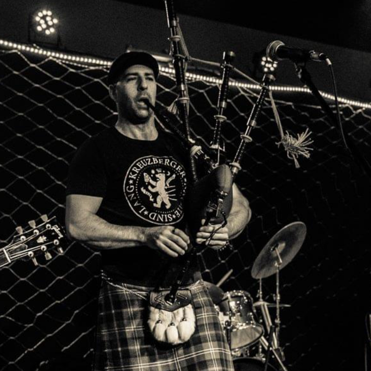 A man playing bagpipes on stage in a black T-shirt and plaid kilt, with a drum set and microphone nearby, in a music venue.