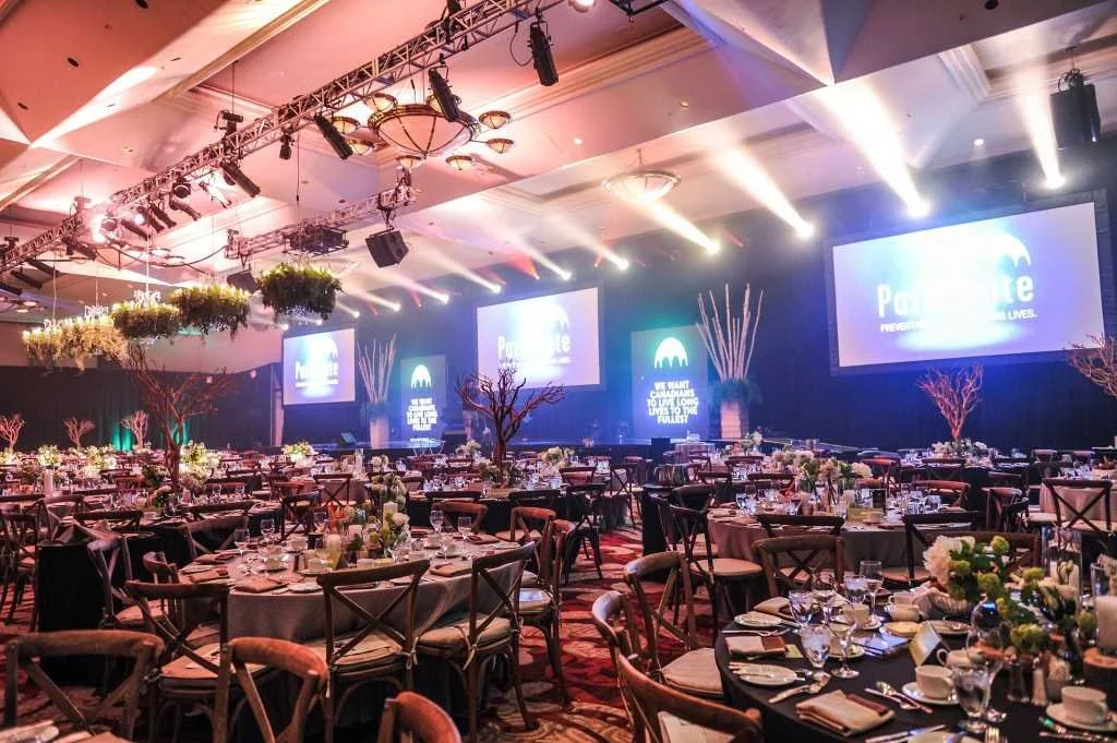 A large event hall decorated with round tables set for a formal dinner, featuring floral centerpieces and place settings. The stage has large screens displaying event branding, with lighting and sound equipment overhead, and decorative trees and hanging plants enhance the elegant atmosphere.