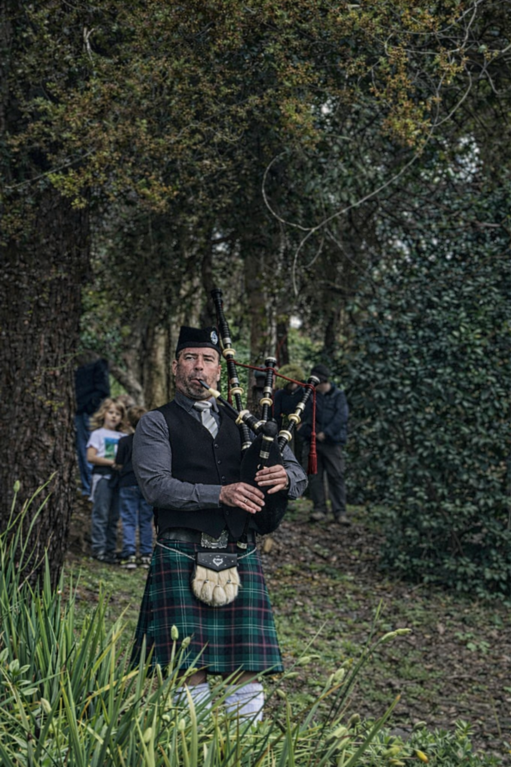 A man dressed in traditional Scottish attire playing the bagpipes outdoors, with a background of trees and a group of children and adults.