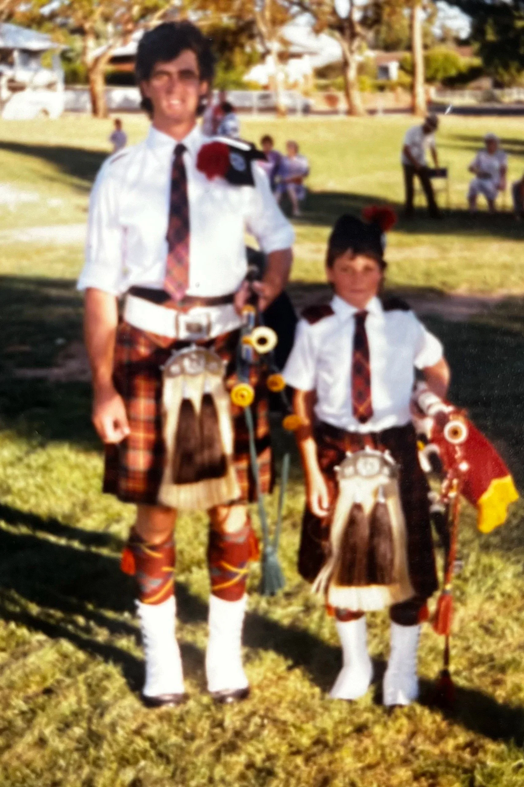 Two boys dressed in traditional Scottish attire, standing outdoors on grass, with others sitting and standing in the background.
