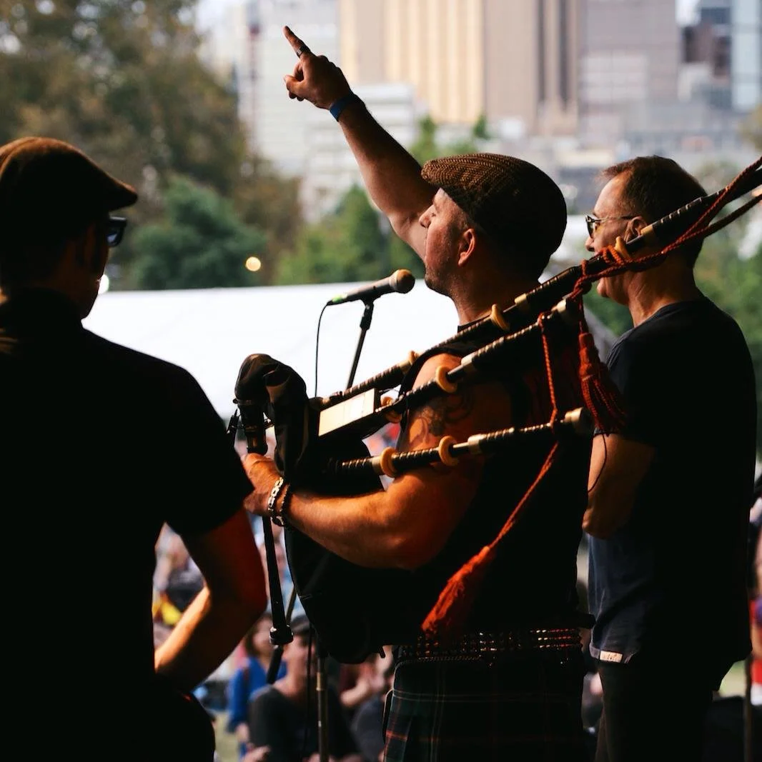 A man playing bagpipes on a stage, raising his right arm with a pointed finger, during a performance outdoors with an audience and city buildings in the background.