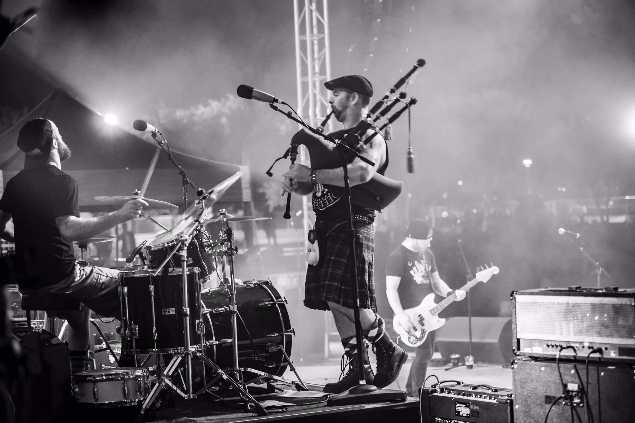 A black-and-white photo of a band performing on stage. The lead musician in the center is playing bagpipes and wearing a kilt. To the left, a drummer is playing the drums, and to the right, a bassist is playing a bass guitar. There are microphones an