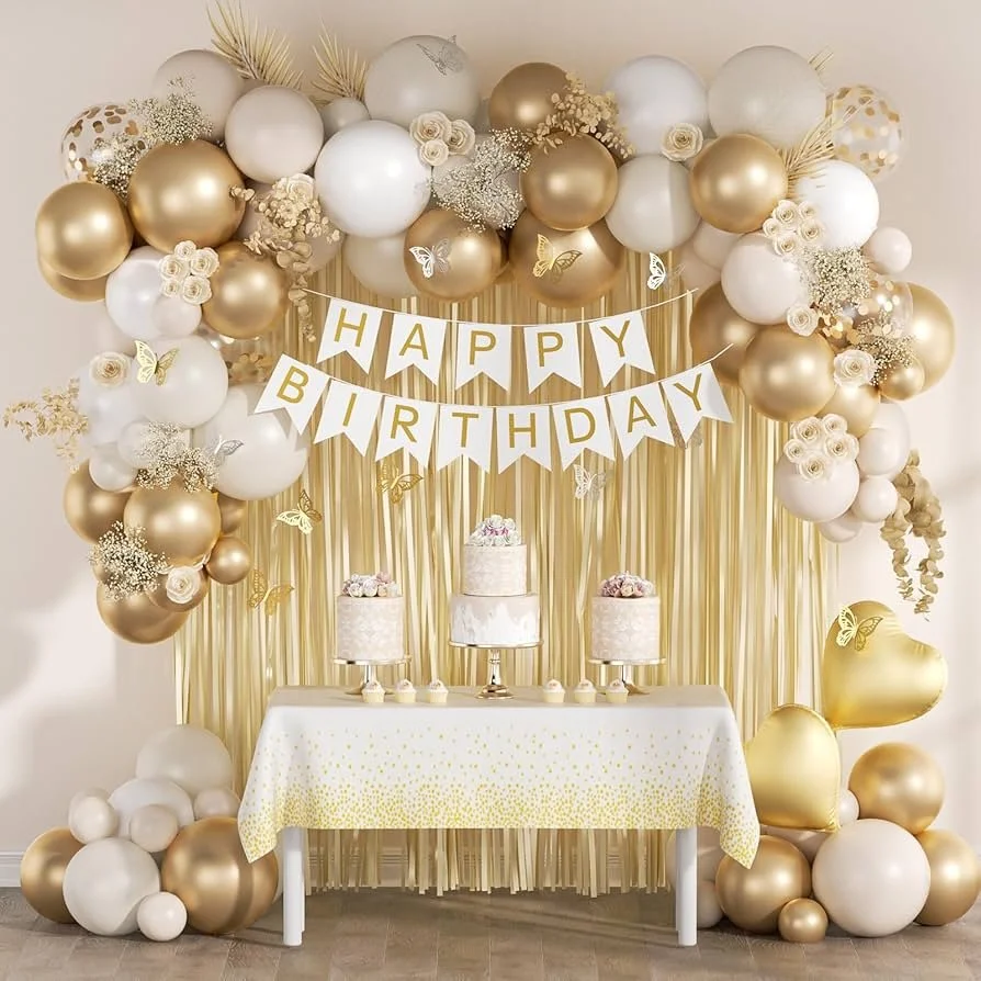 Elegant birthday party setup with gold and white balloons, gold curtains, a 'HAPPY BIRTHDAY' banner, and a table with three decorated cakes and small desserts.