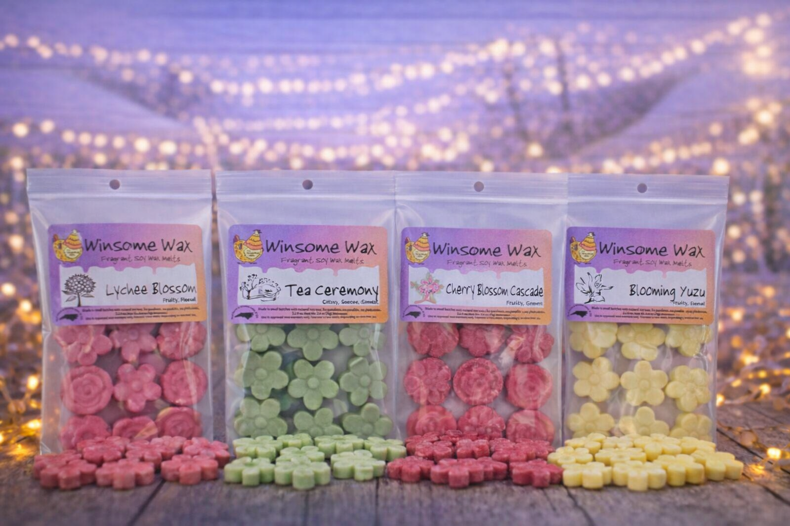 Four packets of Winsome Wax fragrant soy wax melts with flower-shaped wax pieces in pink, green, red, and yellow colors, arranged on a wooden surface against a background of glowing string lights and purple flowers.