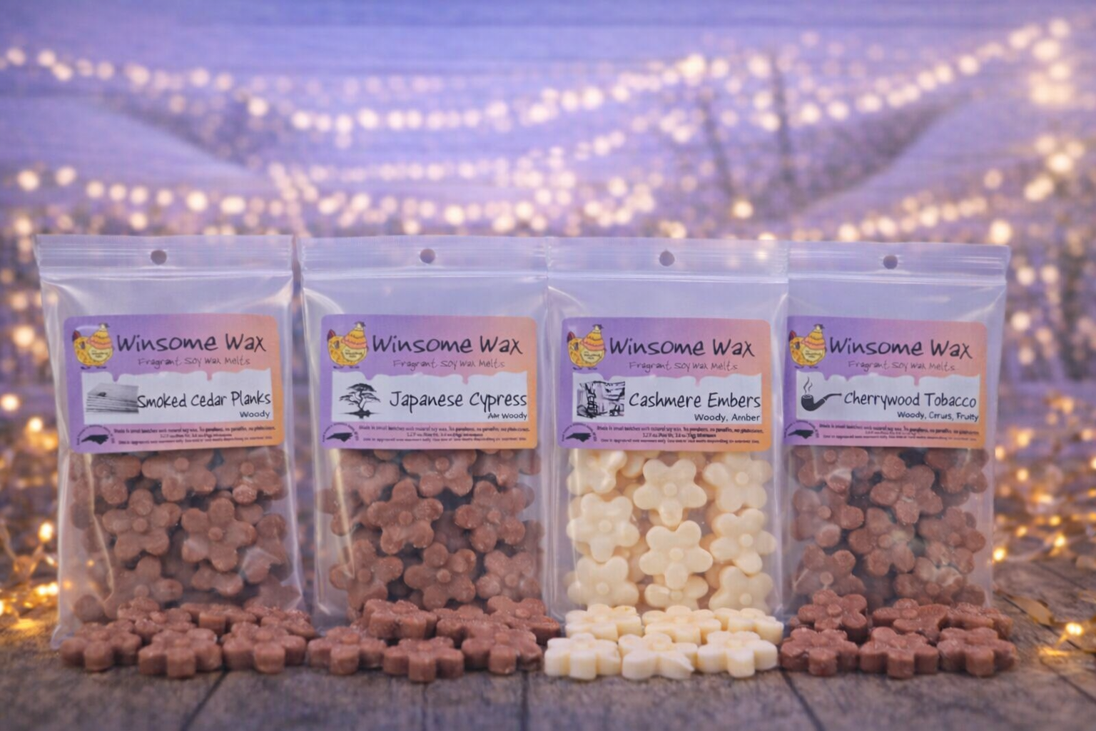 Four small resealable bags of wax melts labeled 'Winsome Wax' with different scents, including smoked cedar planks, Japanese cypress, cashmere embers, and cherrywood tobacco, displayed on a wooden surface with soft fairy lights in the background.