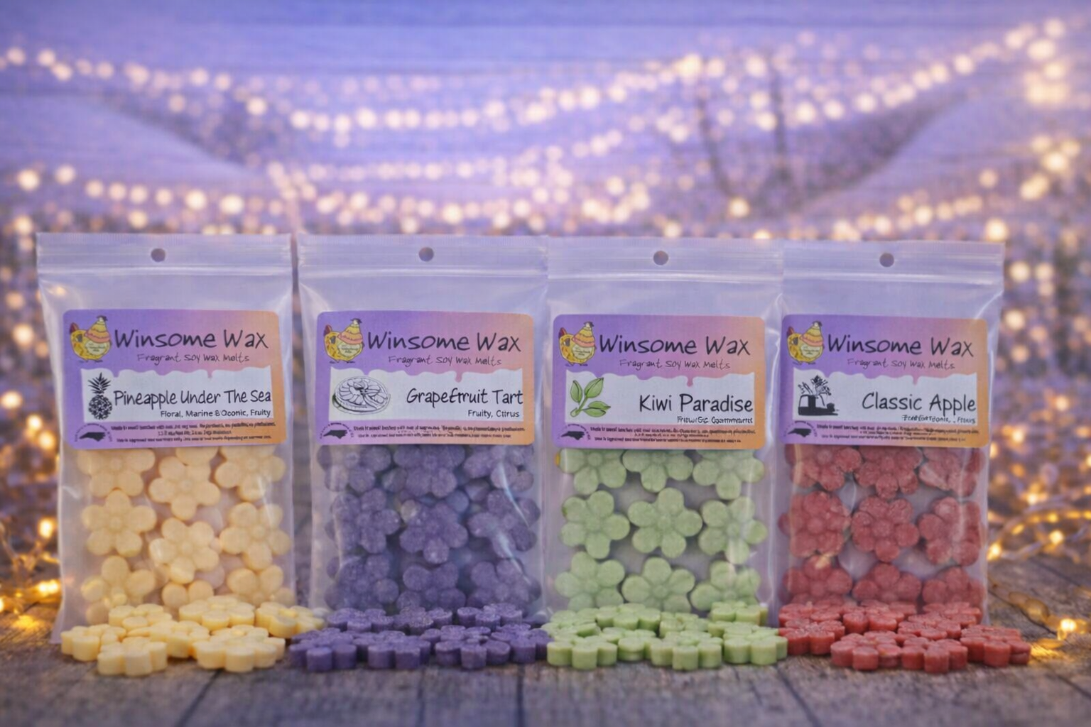 Four clear plastic bags of scented wax melts with labels displaying the scents: Pineapple Under The Sea, Grapefruit Tart, Kiwi Paradise, and Classic Apple, arranged on a wooden surface with a blurred background of fairy lights.