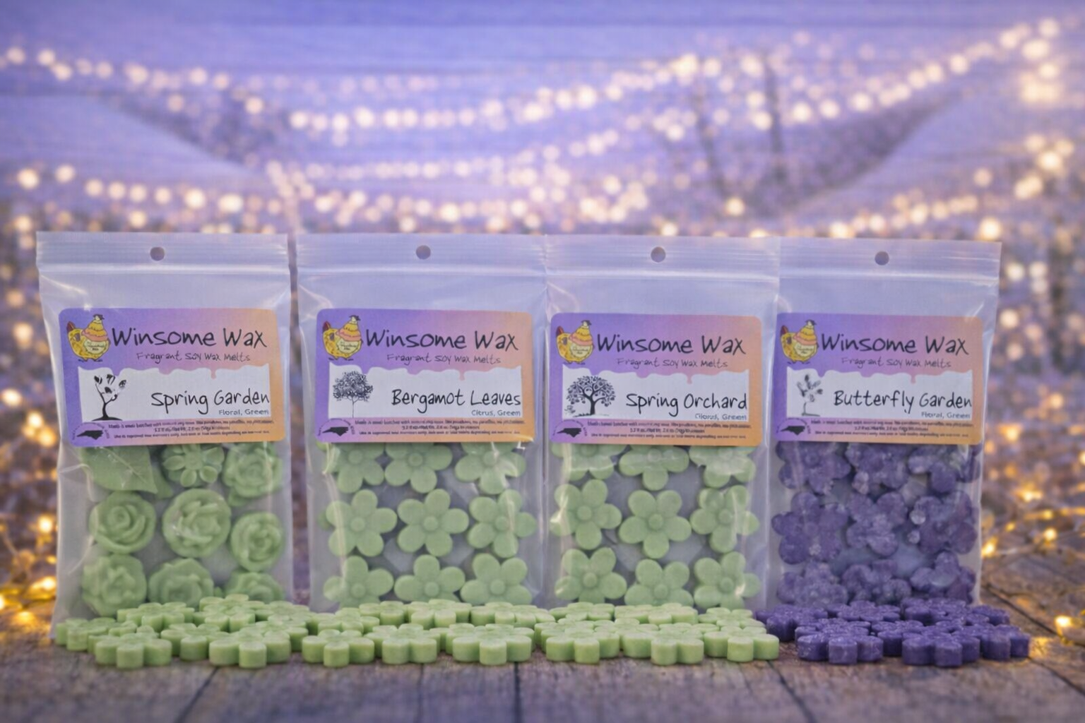 Four clear plastic bags of scented soy wax melts with spring-themed labels, arranged in a row on a wooden surface with fairy lights in the background. The scents are Spring Garden, Bergamot Leaves, Spring Orchard, and Butterfly Garden.