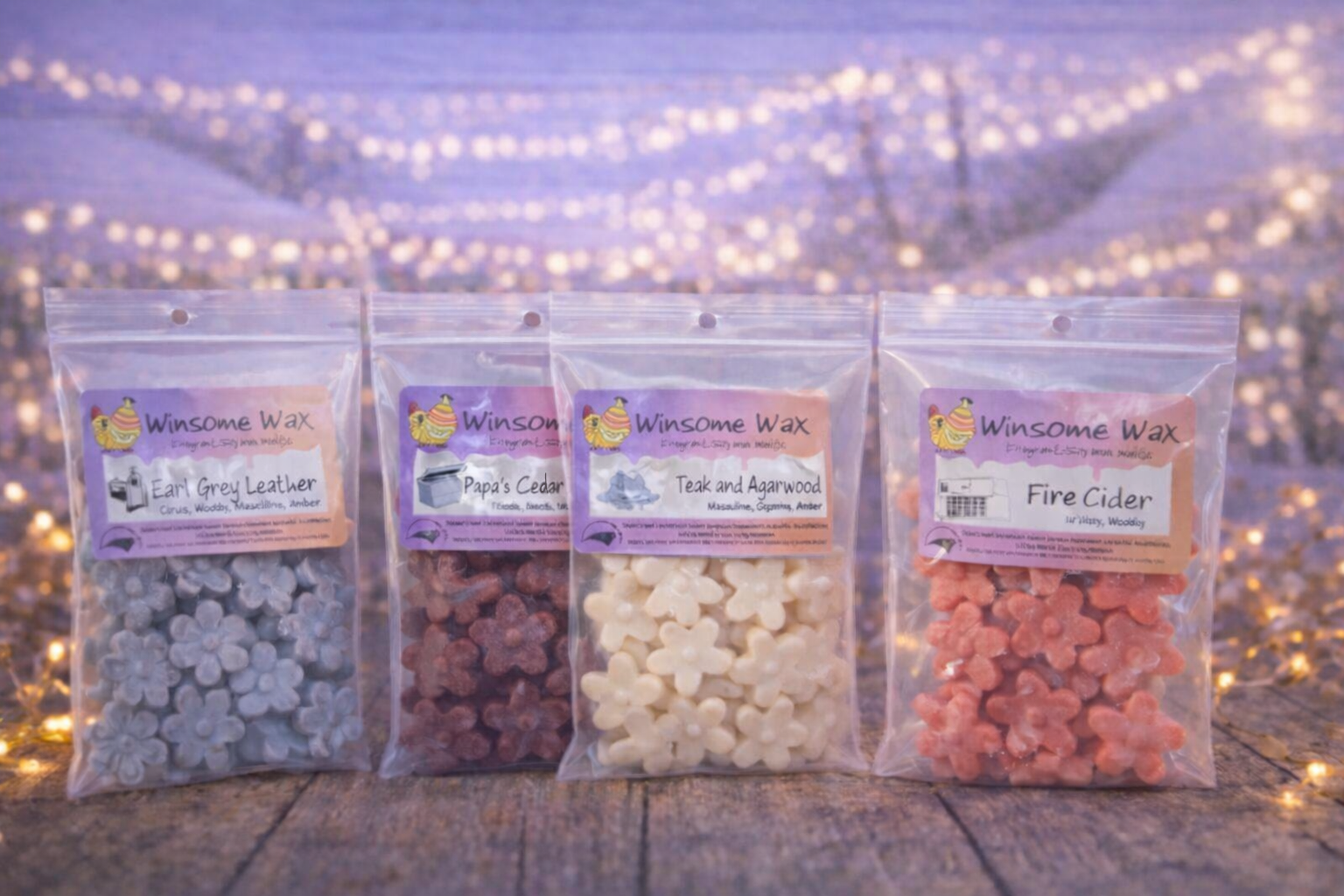 Four clear plastic bags of scented wax melts with floral shapes, labeled with different scents, placed on a wooden surface with fairy lights in the background.