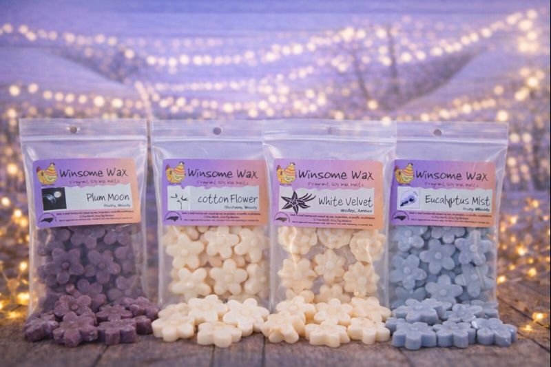 Four resealable bags of wax melts labeled with different scents, including Plum Moon, Cotton Flower, White Velvet, and Eucalyptus Mist, displayed on a wooden surface with string lights in the background.