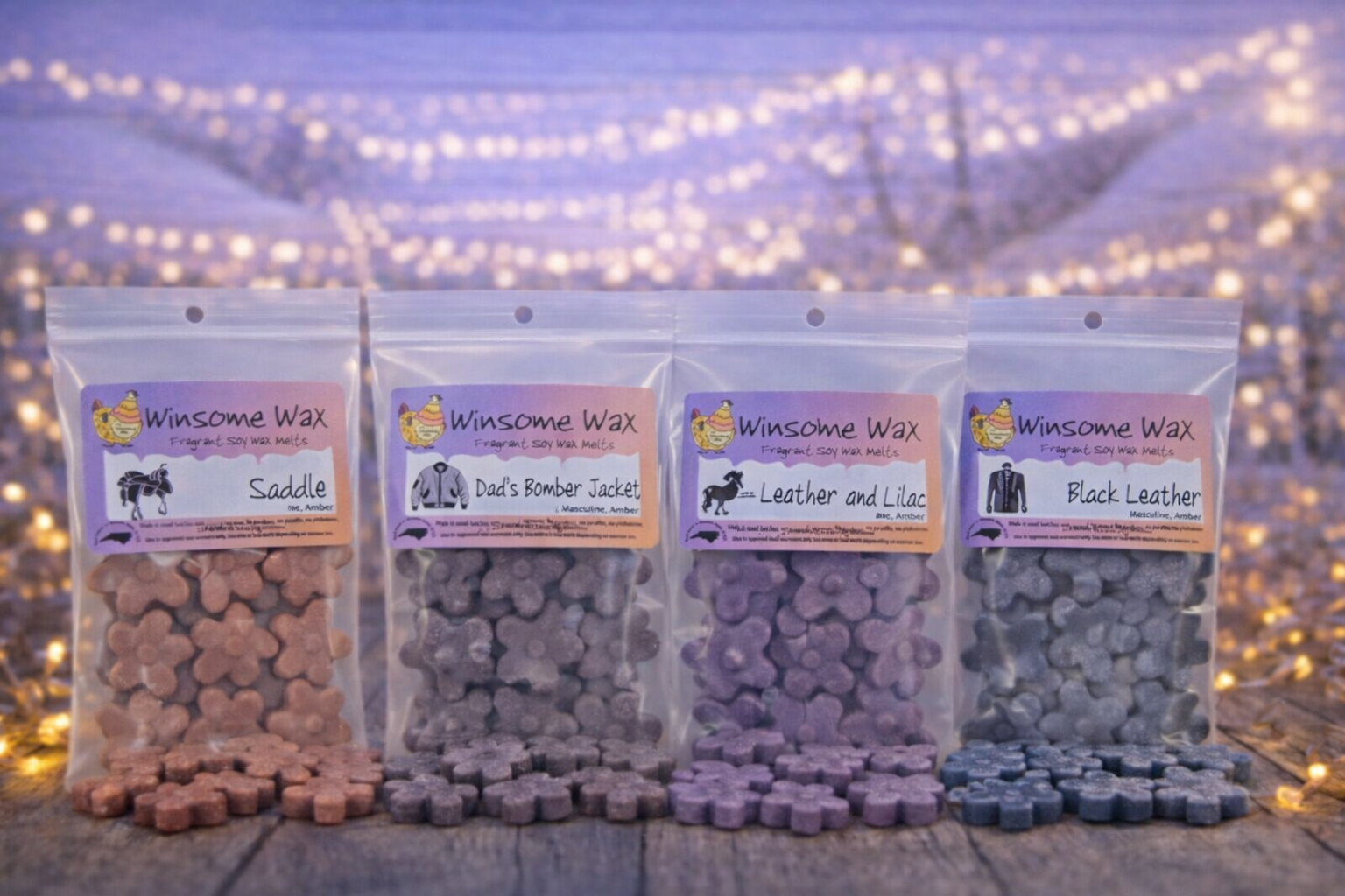 Four bags of Winsome Wax fragrant soy wax melts in animal-shaped hearts, arranged on a wooden surface with a blurred string of fairy lights in the background. The scents include Saddle, Dad's Bomber Jacket, Leather and Lilac, and Black Leather.