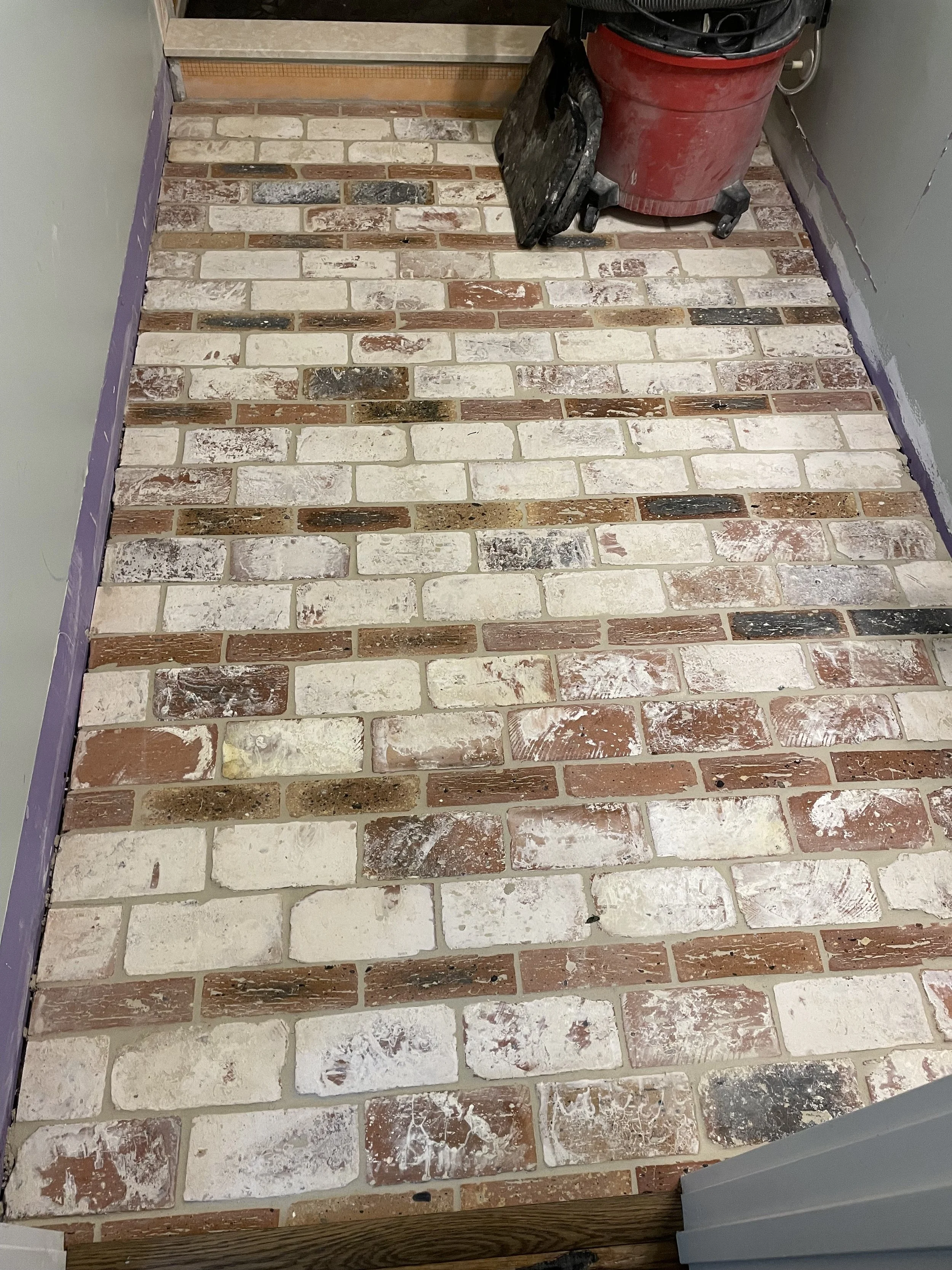 Brick floor with various white, tan, red, and black bricks, some with paint or stain marks, near a wall with purple painter's tape on the edges, and a red vacuum cleaner with a black surface next to it.
