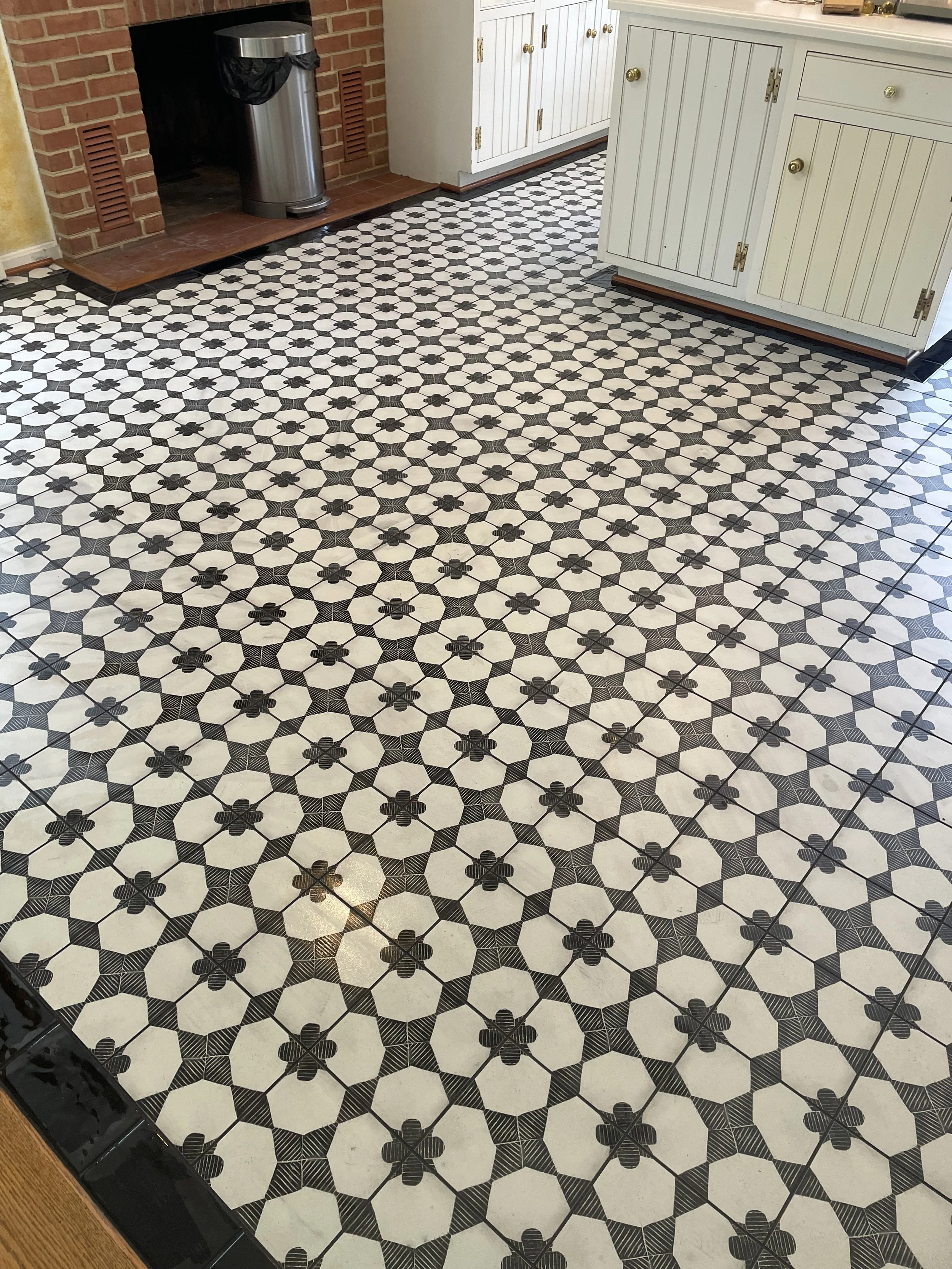 A tiled kitchen floor with black and white geometric and floral patterns, with white cabinets and brick fireplace in the background.