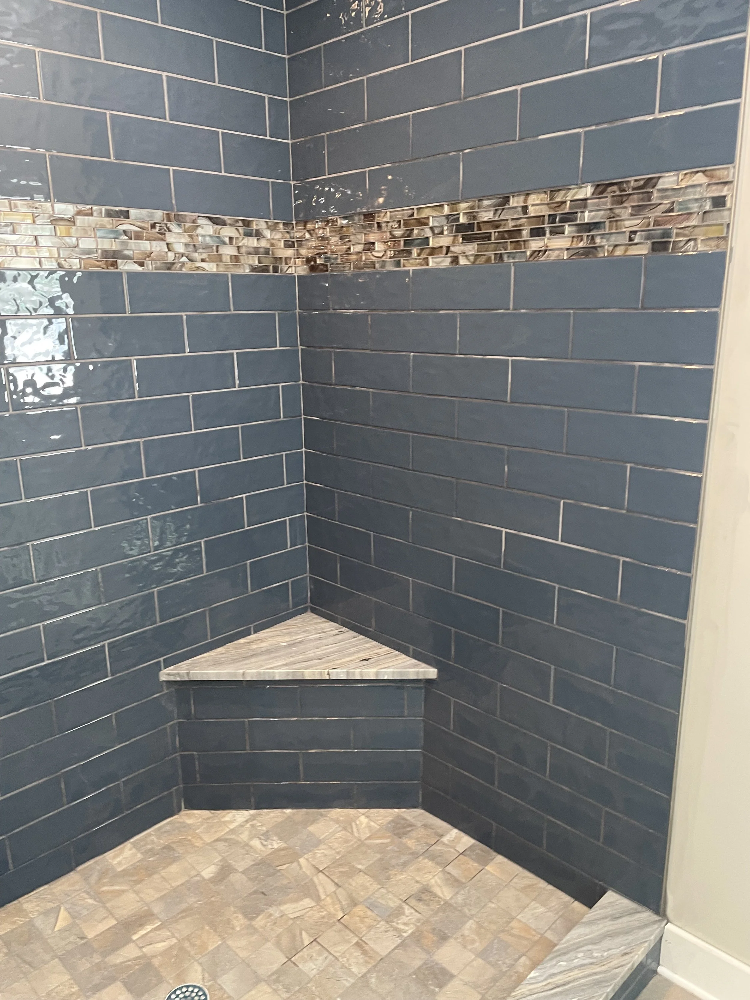Corner shower with navy blue subway tiles, a strip of multicolored small mosaic tiles, a marble corner bench, and a mosaic tiled floor.