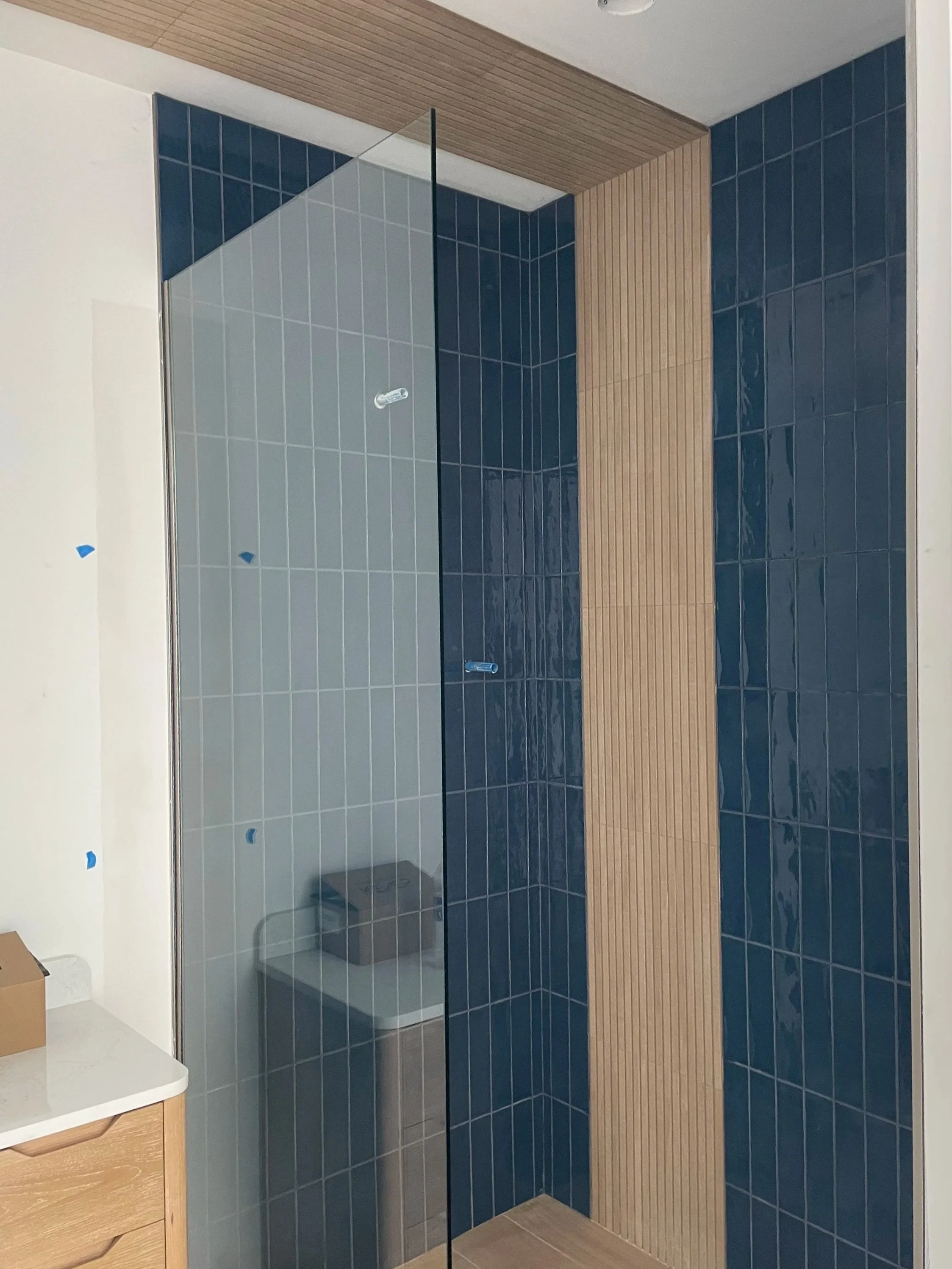 A partially finished walk-in shower with dark blue tiles, a glass door, and light wooden accents.