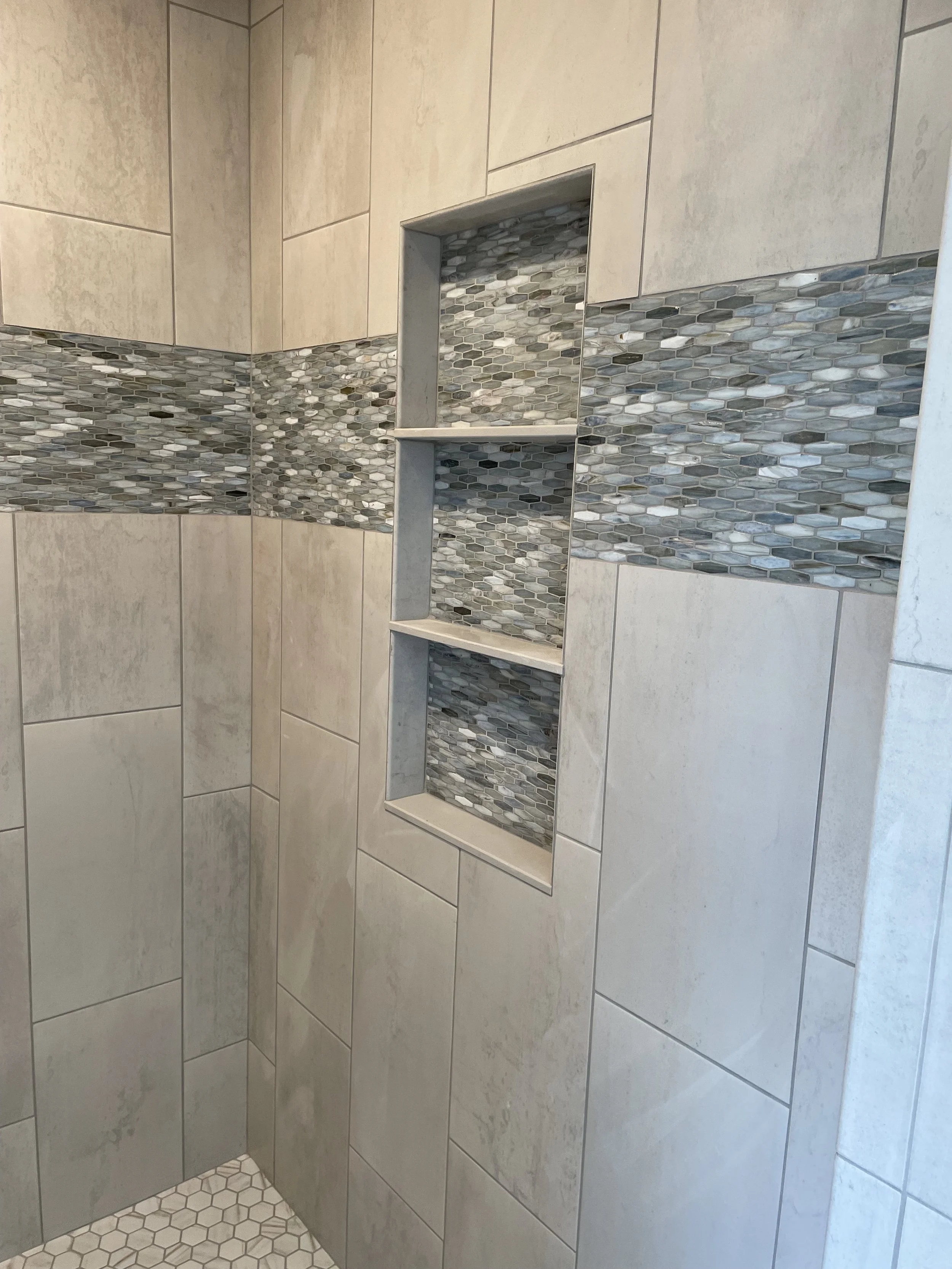 A tiled shower wall featuring large beige tiles, a horizontal band of small gray and white mosaic tiles, and a built-in niche with three shelves lined with matching mosaic tiles.