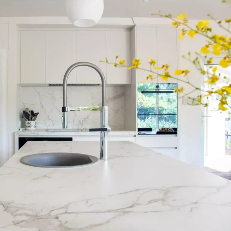 Modern kitchen with white marble countertops, a round sink, and a tall curved faucet, with yellow flowering branches in the foreground.