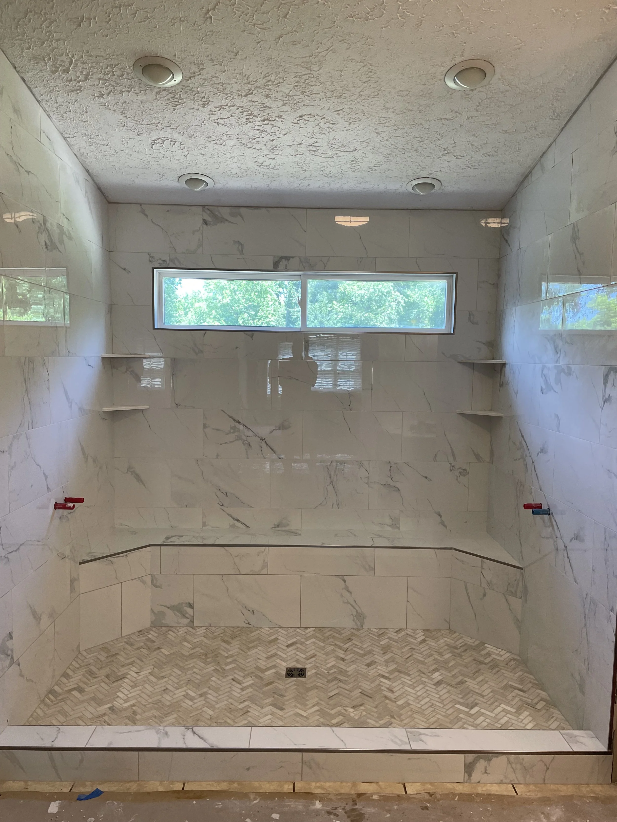 A bathroom shower with marble tiles on the walls, a small horizontal window, and built-in corner shelves, with no shower fixtures installed yet.