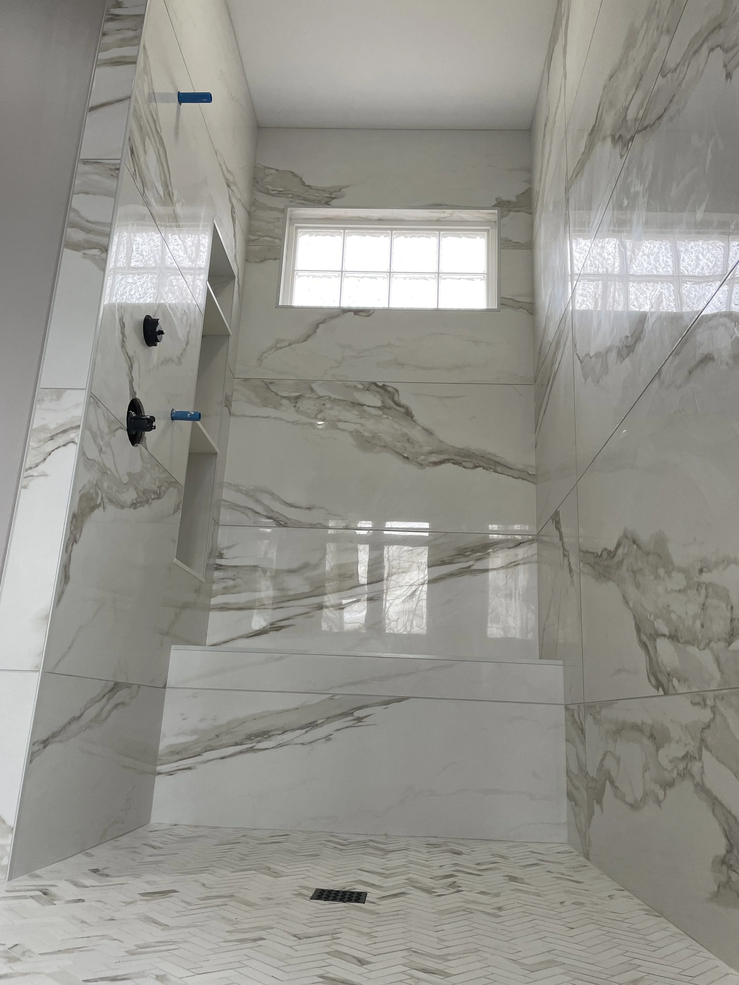 Modern marble-tiled walk-in shower with a high window, built-in niche, and a drain on the floor.