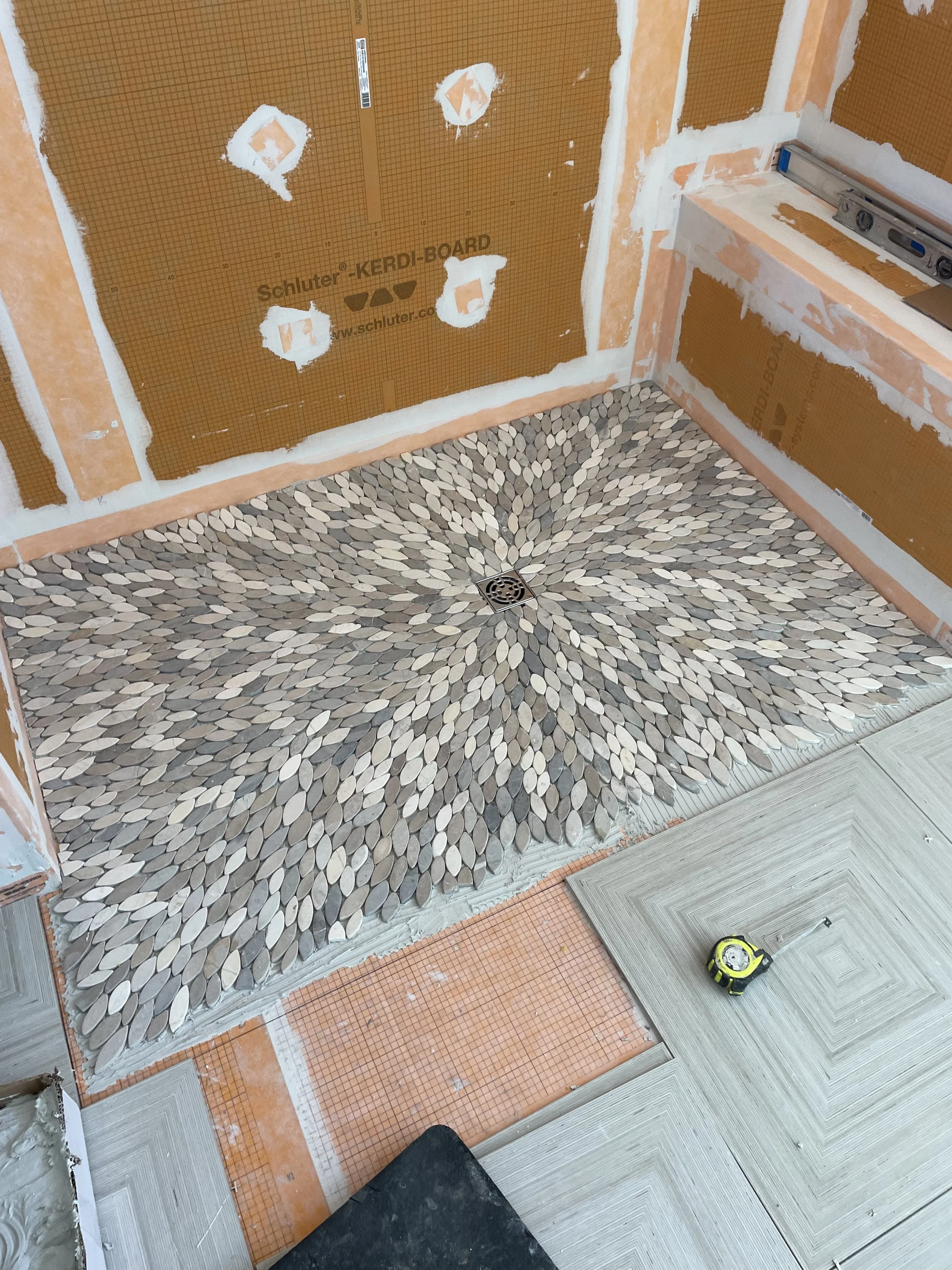 A partially tiled bathroom floor with an intricate pebble mosaic pattern in a shower floor, a level tool, and unfinished wall tiling.