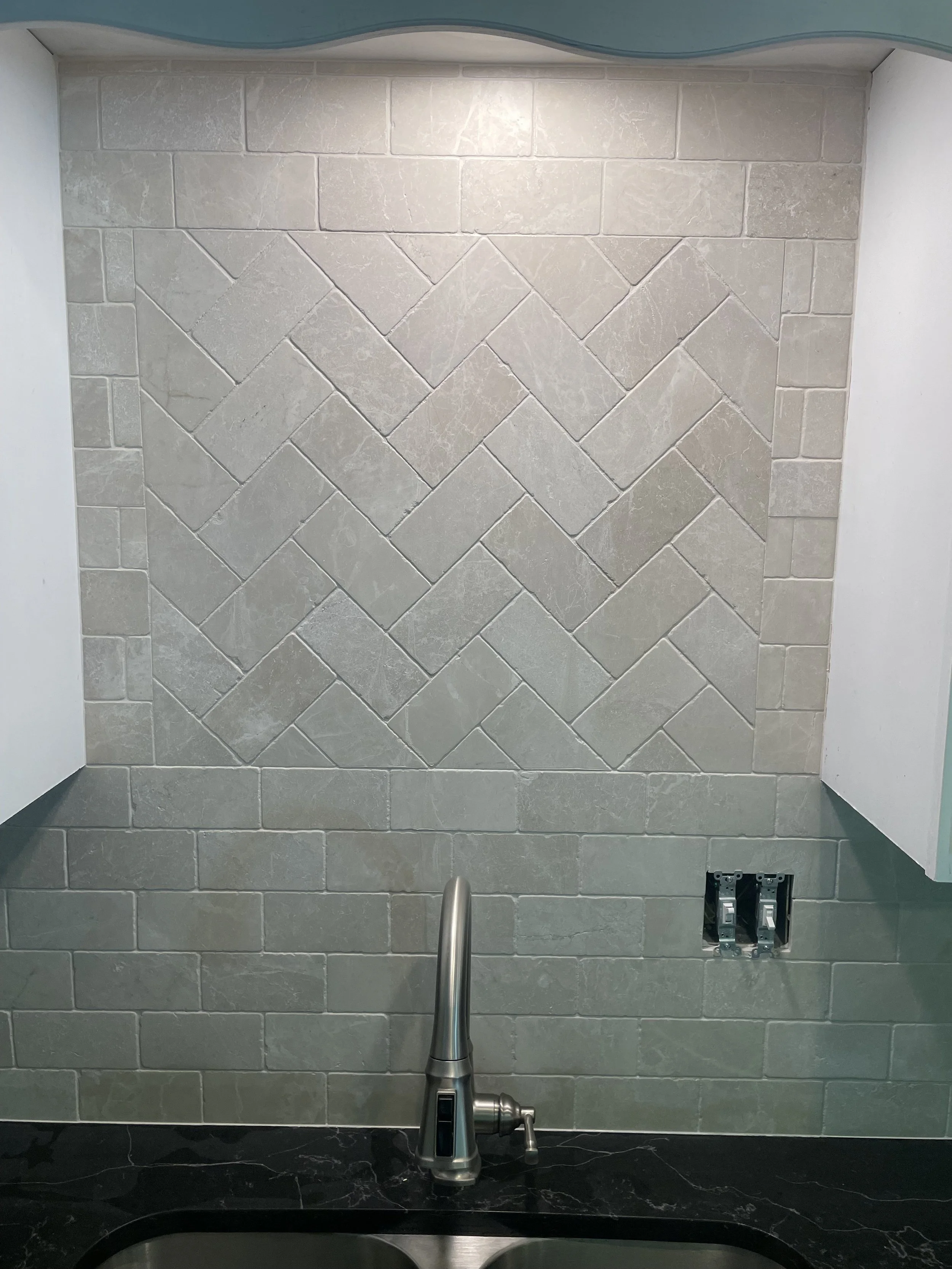 Kitchen backsplash with beige stones arranged in a herringbone pattern. There is a faucet in the foreground above a black countertop, and an electrical outlet missing cover on the right side.