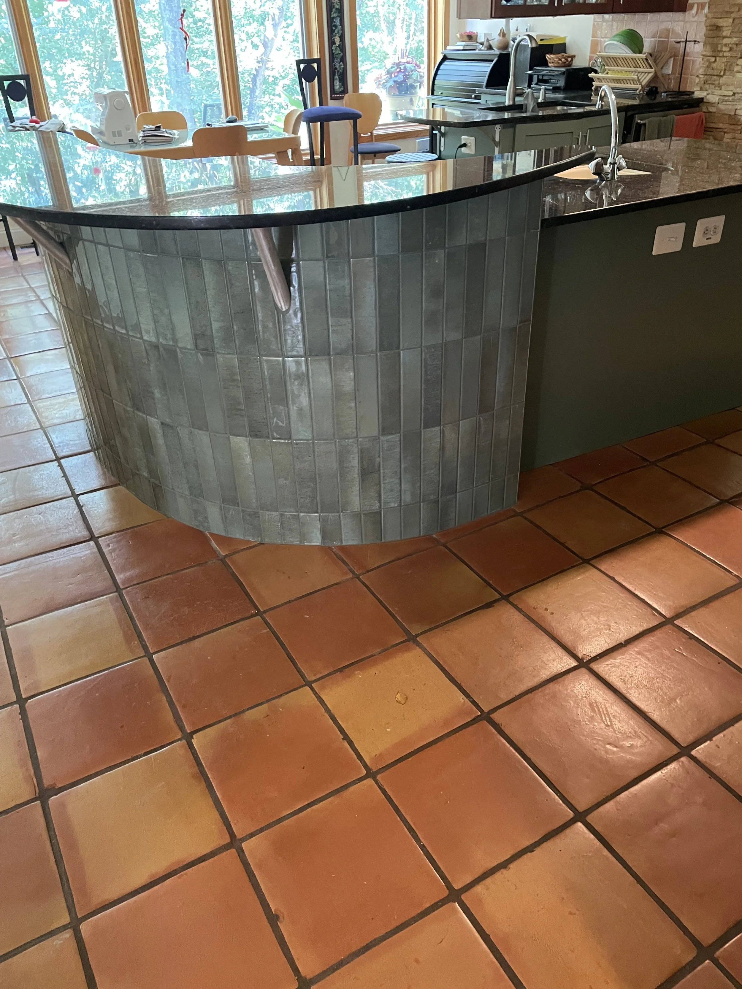 Close-up of a kitchen island with a curved edge, covered in dark granite, in a kitchen with terracotta floor tiles and gray-green tiled base. In the background, there are wooden chairs around a dining table near large windows, with kitchen appliances