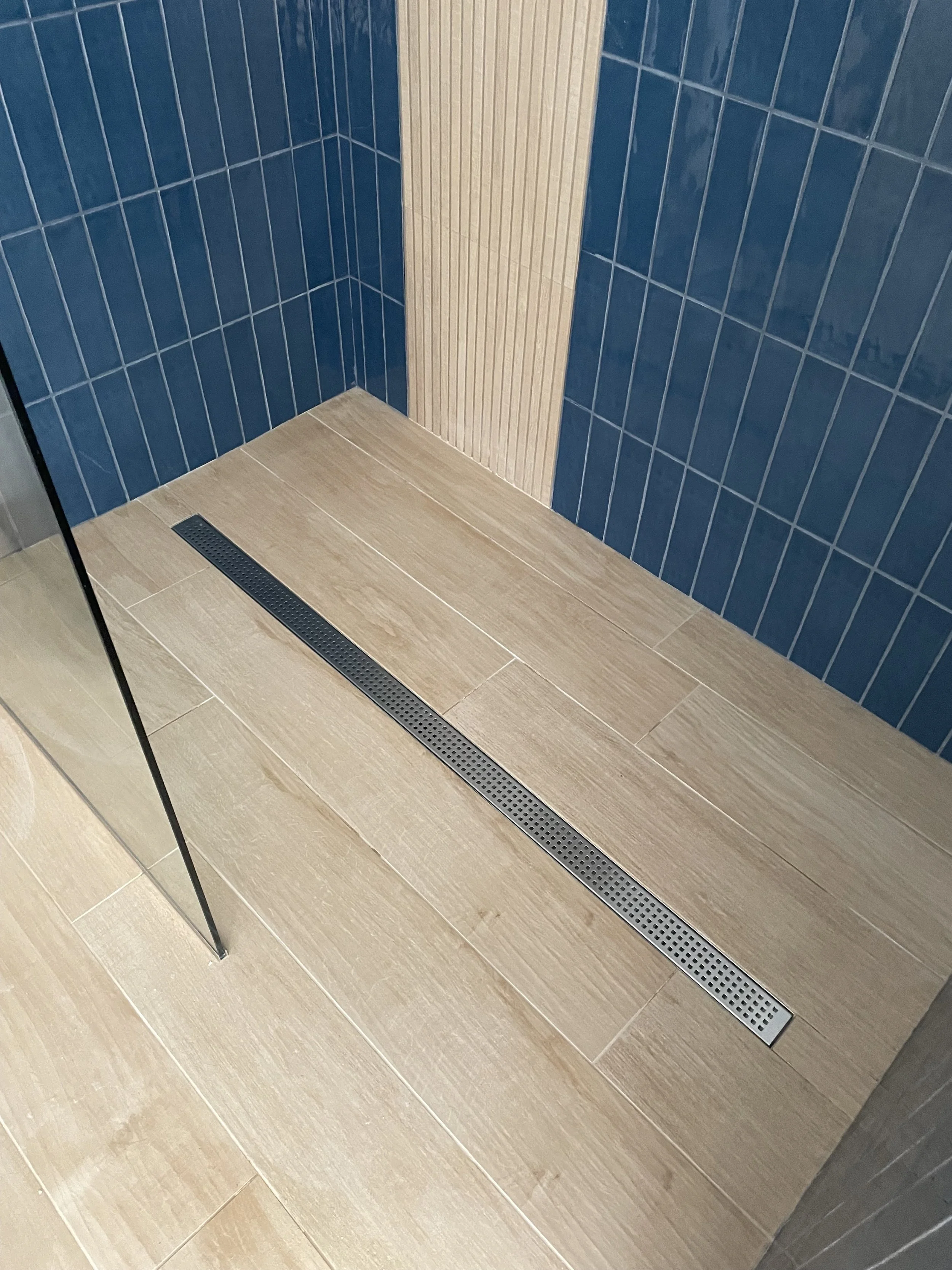 Close-up of a shower floor with a linear drain, blue and beige tiled walls, and a glass shower partition.