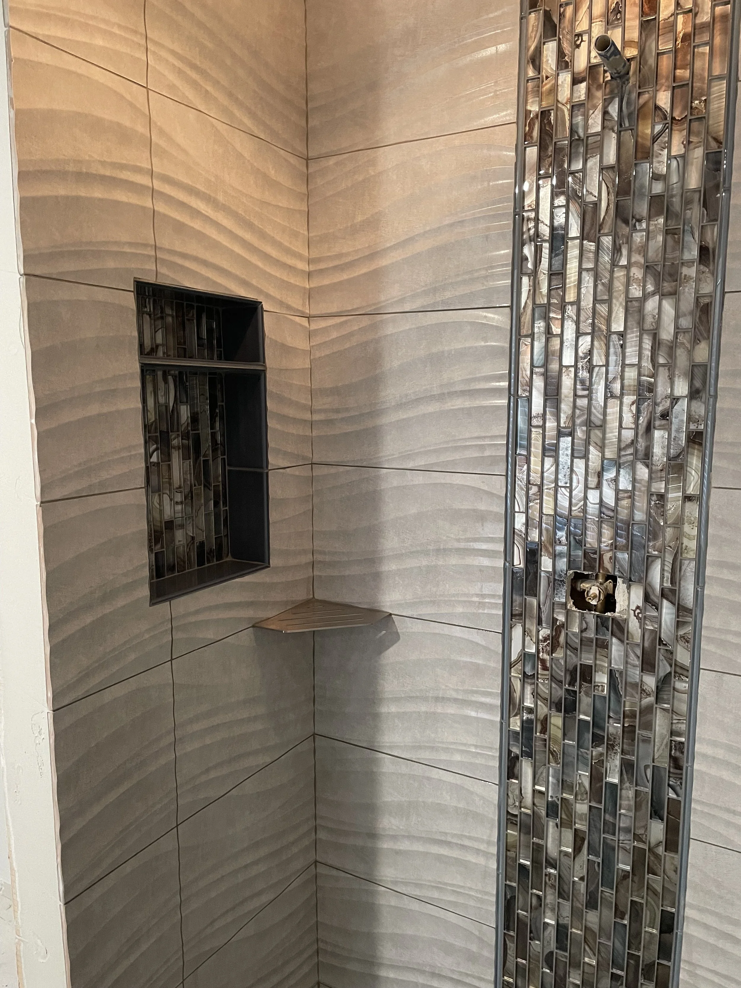 Empty shower with beige textured wall tiles, a built-in black tile niche, and a vertical strip of metallic mosaic tiles.