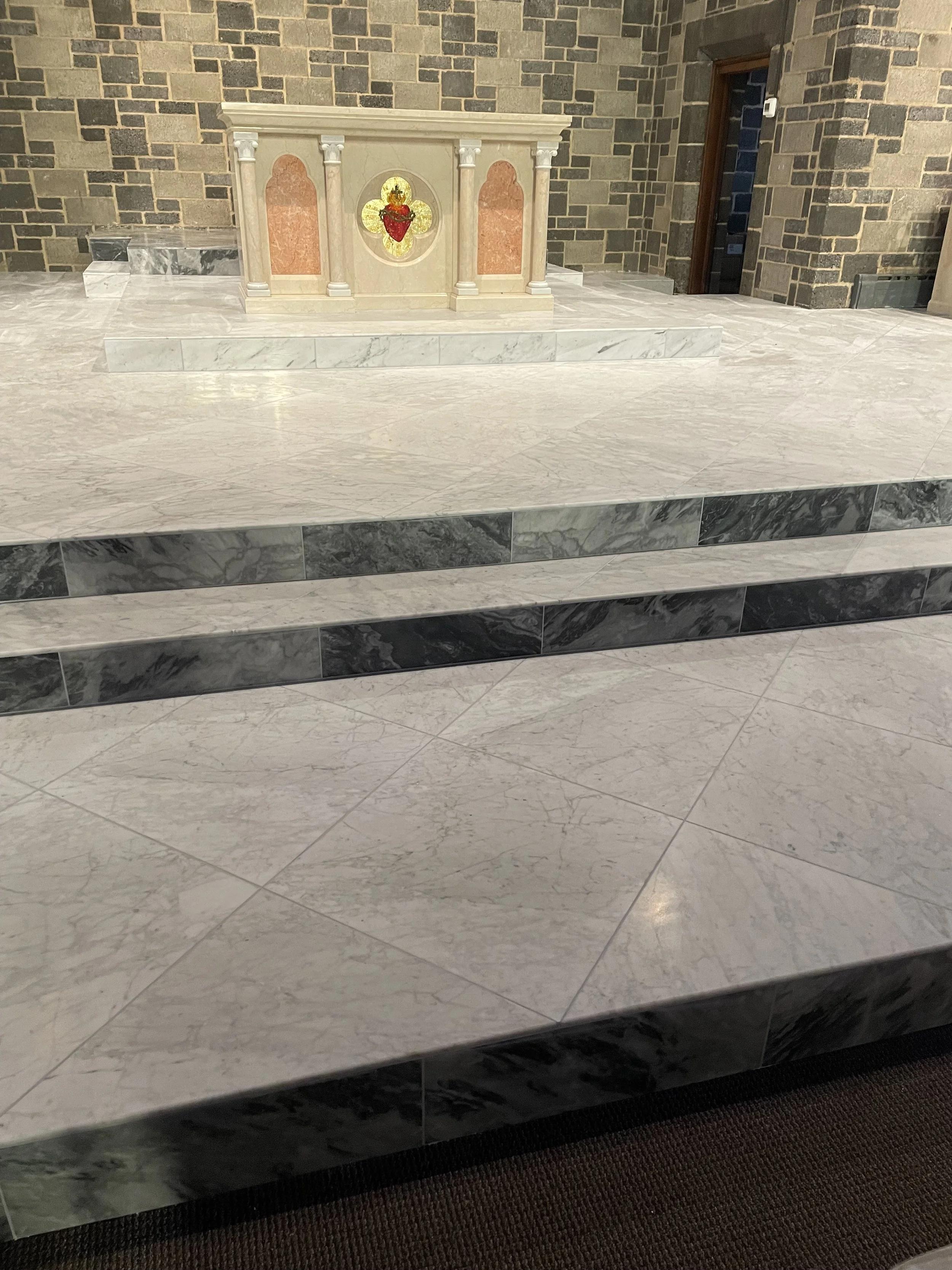 An altar in a church with marble steps, a decorative marble backdrop, and a small decorated altar table with a red heart and gold cross.