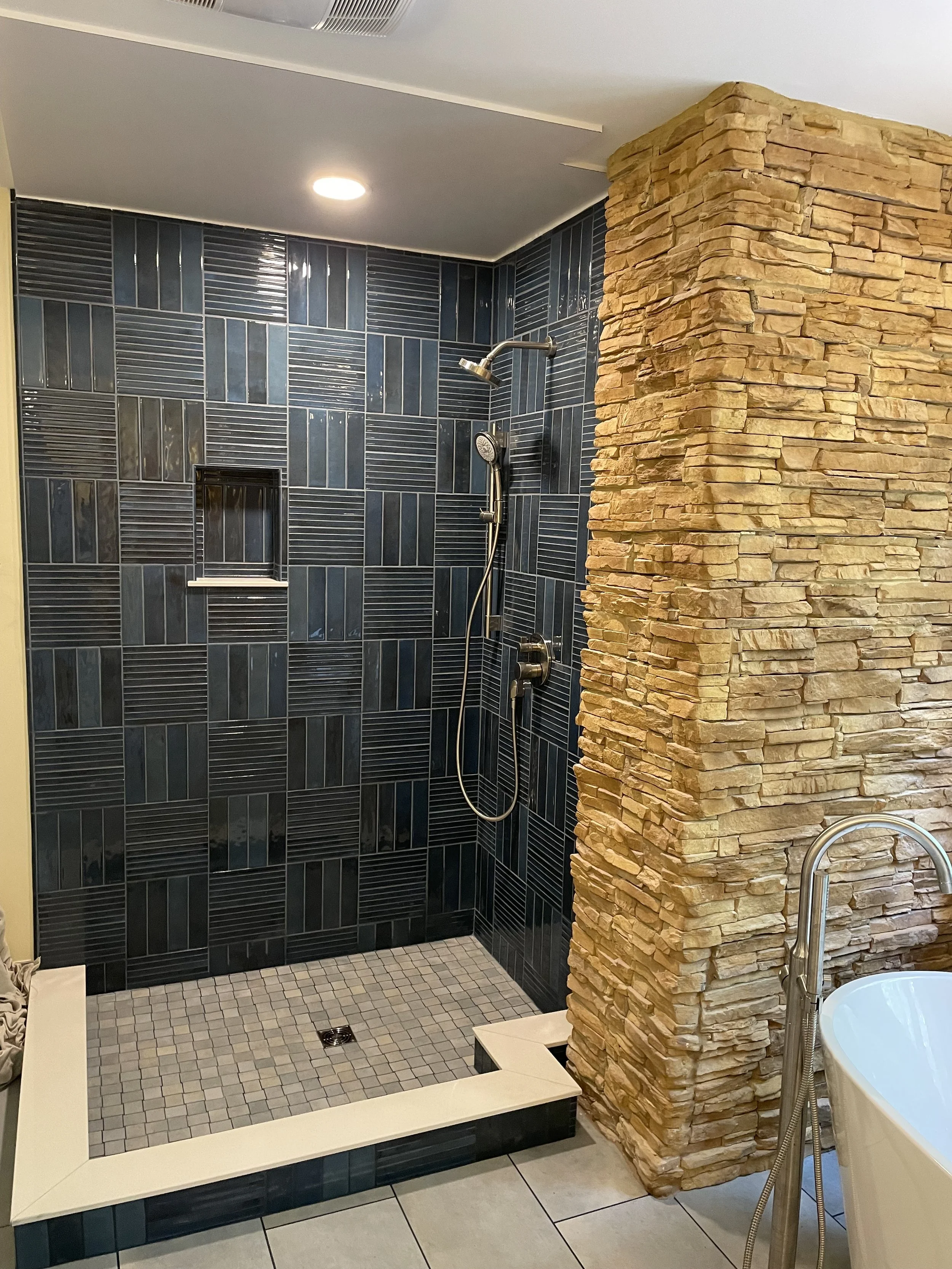 A modern bathroom shower with black tiled walls, featuring a built-in shelf, and fitted with a rainfall and handheld showerhead, separated by a rustic stone wall, with a partial view of a bathtub with a freestanding faucet.