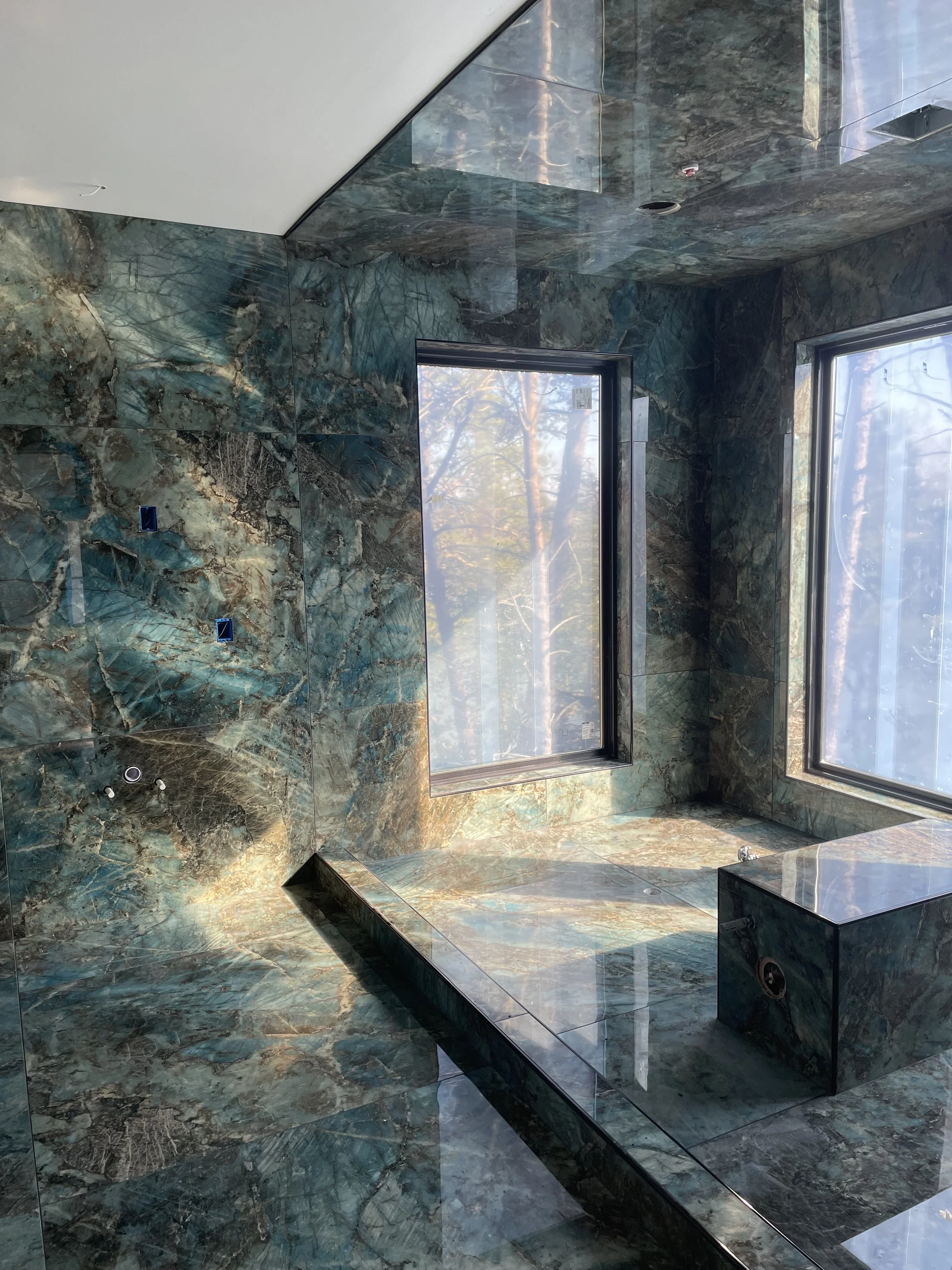 A modern bathroom with marble walls and floor, large windows, and a built-in bench seat.