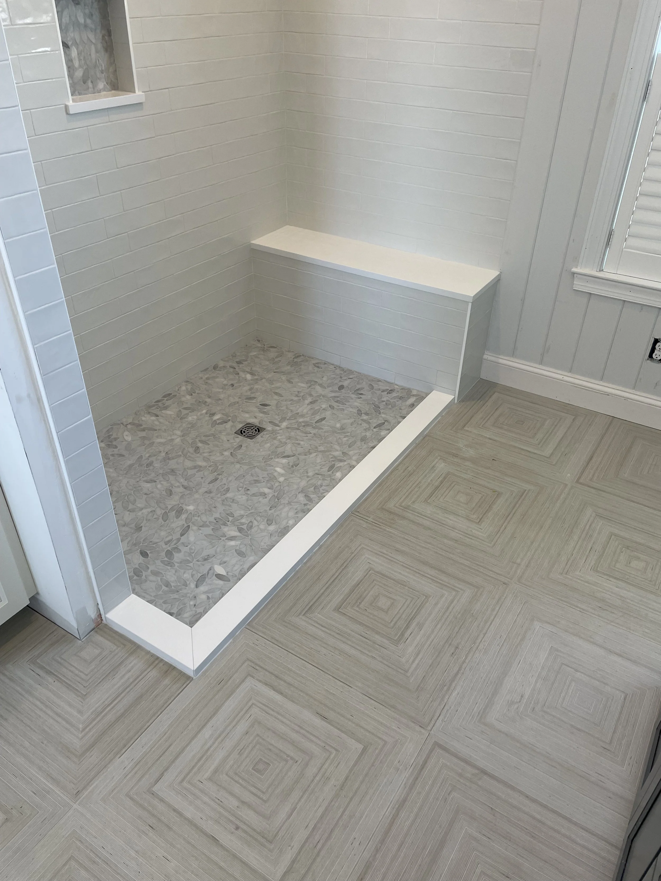 Empty shower stall with leaf-patterned tile floor, white tiled walls, built-in bench, and a small window with frosted glass.