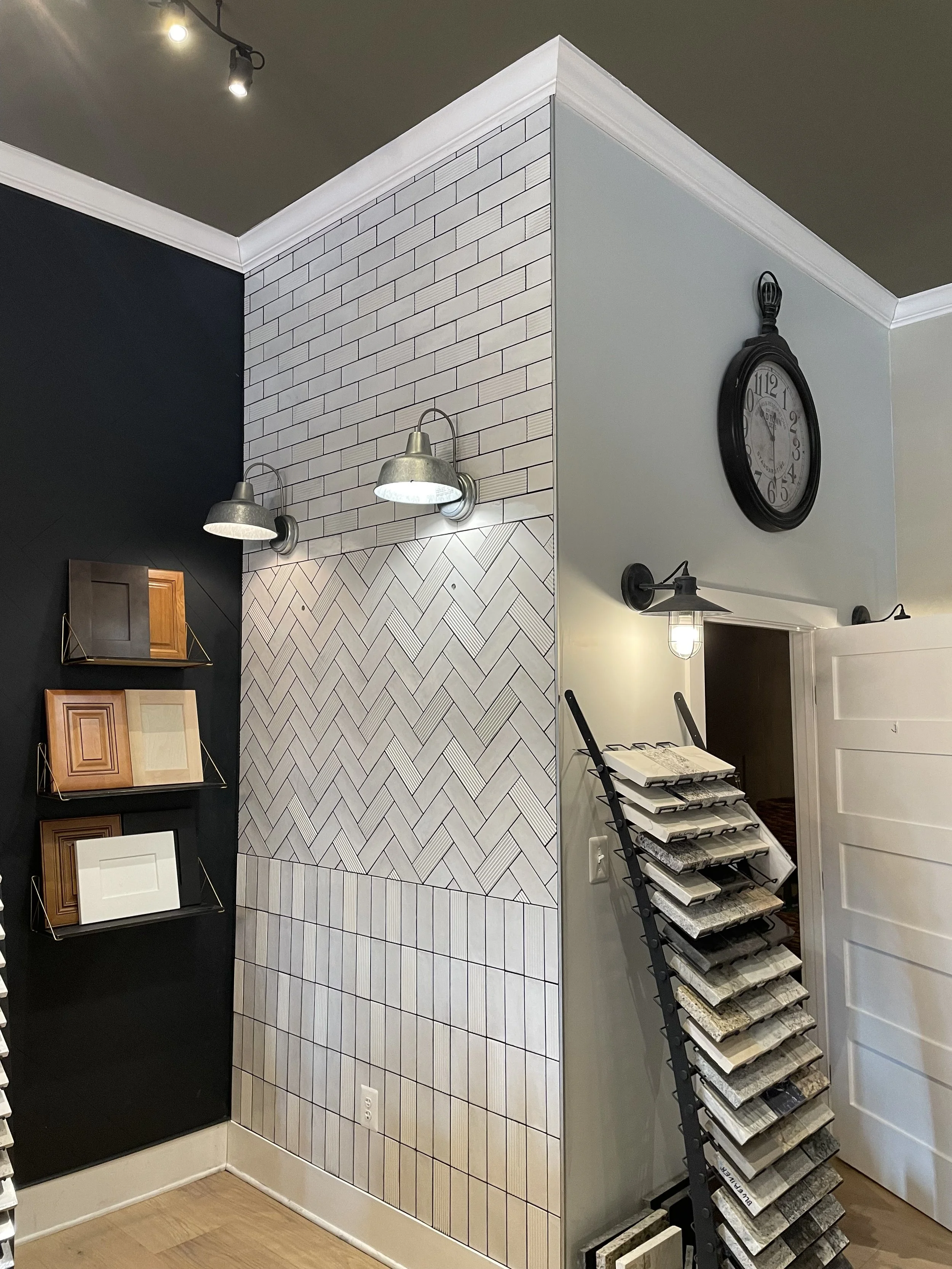 Interior corner of a showroom with decorative wall tiles, picture samples, a large wall clock, and a display rack of tile samples.