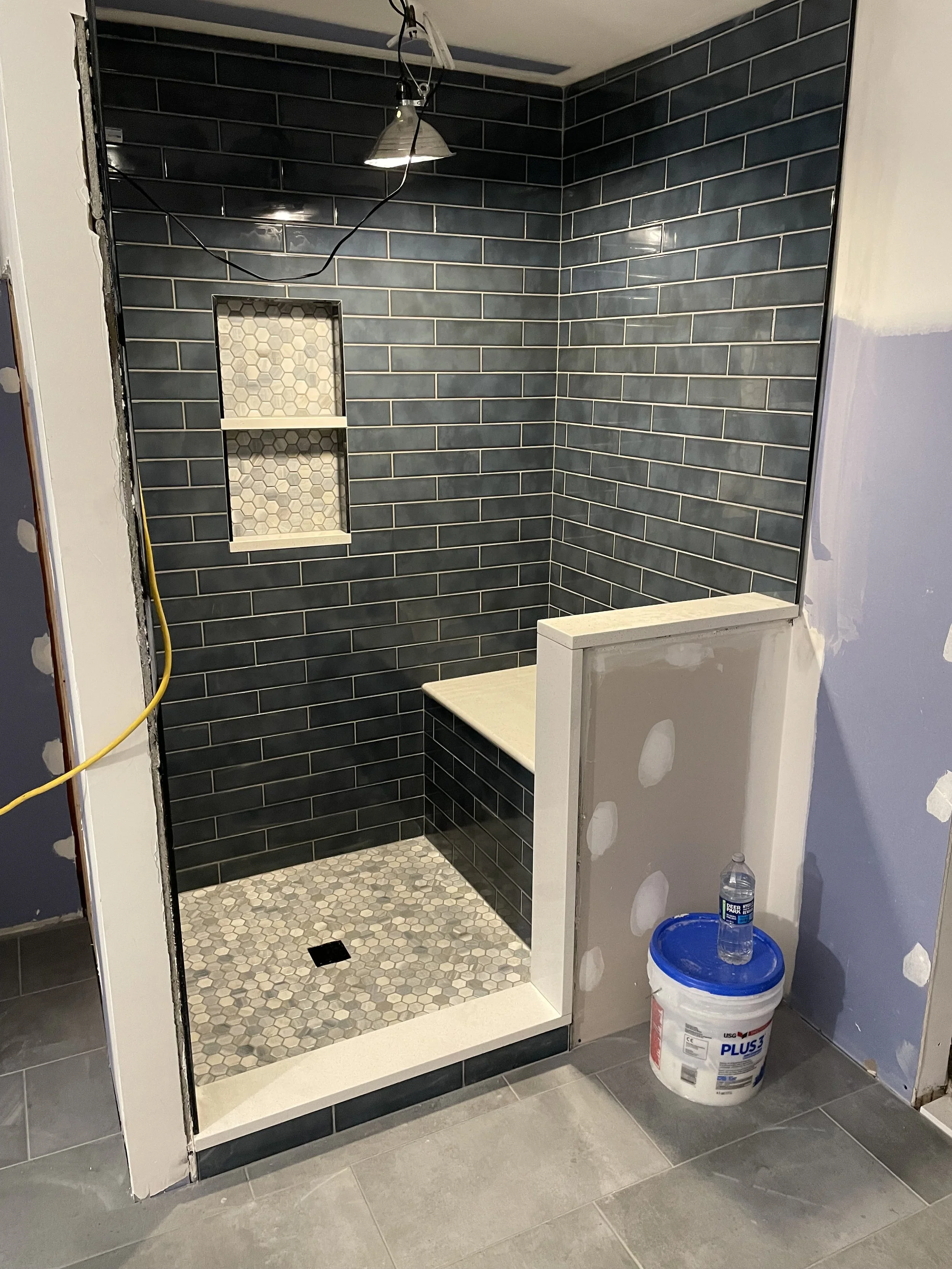 Shower area with dark blue subway tiles, a built-in shelf with hexagon tiles, and a bench with mosaic tiles on the floor. There's a partial partition wall and construction materials on the floor, including a bucket of grout and a water bottle.