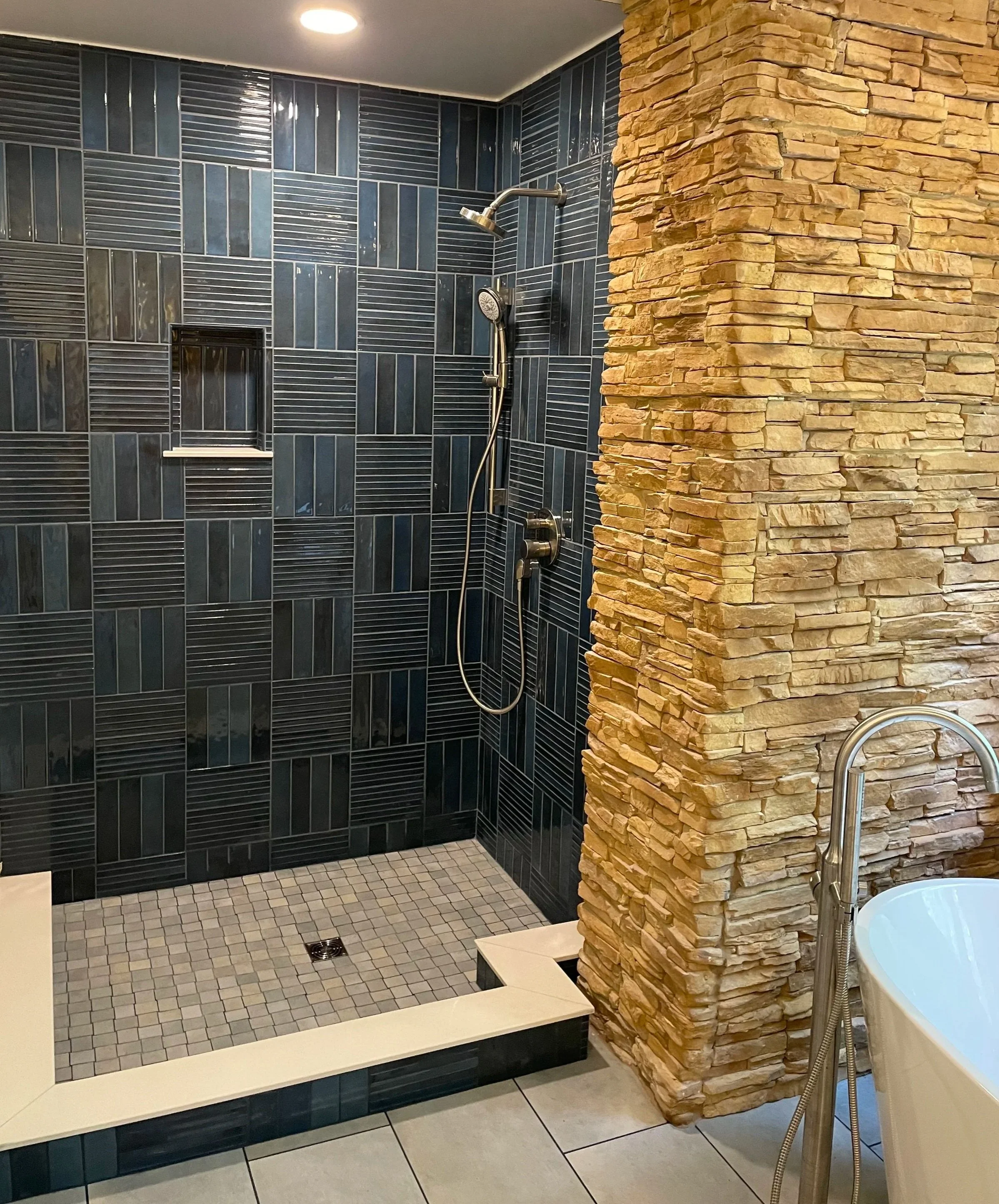 Bathroom shower area with black tiled walls, a built-in shelf, a handheld showerhead, and separate overhead shower, adjacent to a stone accent wall, with a bathtub partially visible on the right.