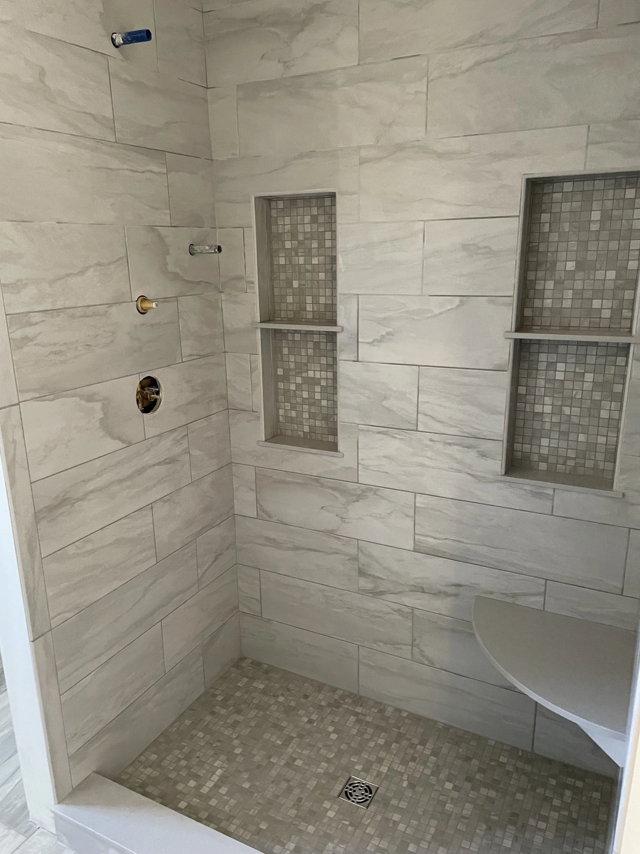 A shower with tiled walls, two recessed shelves with mosaic tiles, a small corner shelf, a drain, and three unfinished shower fixture connections.
