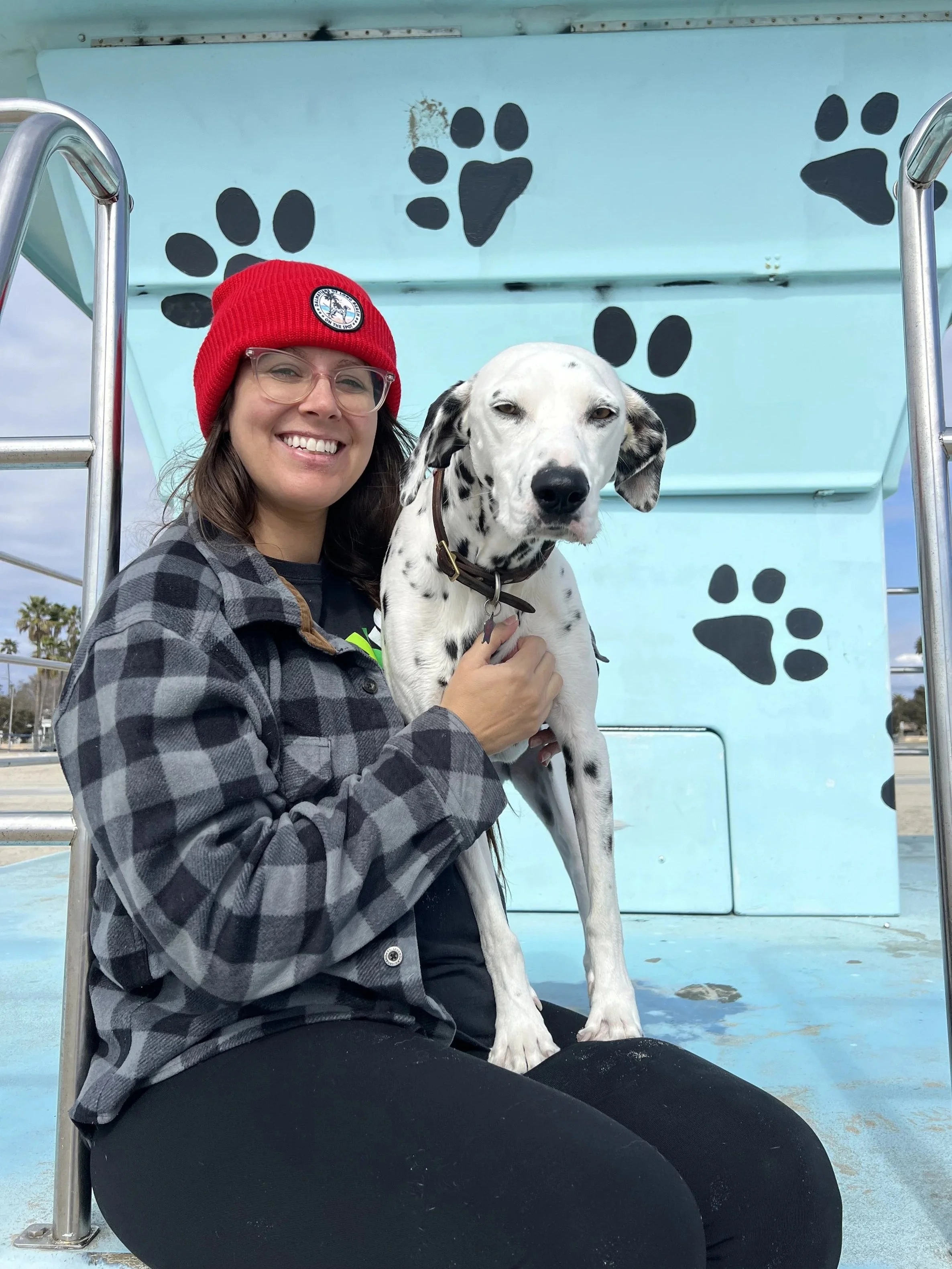 A smiling woman wearing glasses, a red beanie, and a black and gray checkered jacket is sitting on the ground holding a Dalmatian dog with black spots on a blue structure with black paw prints painted on it. The woman is smiling, and the dog has a calm expression.