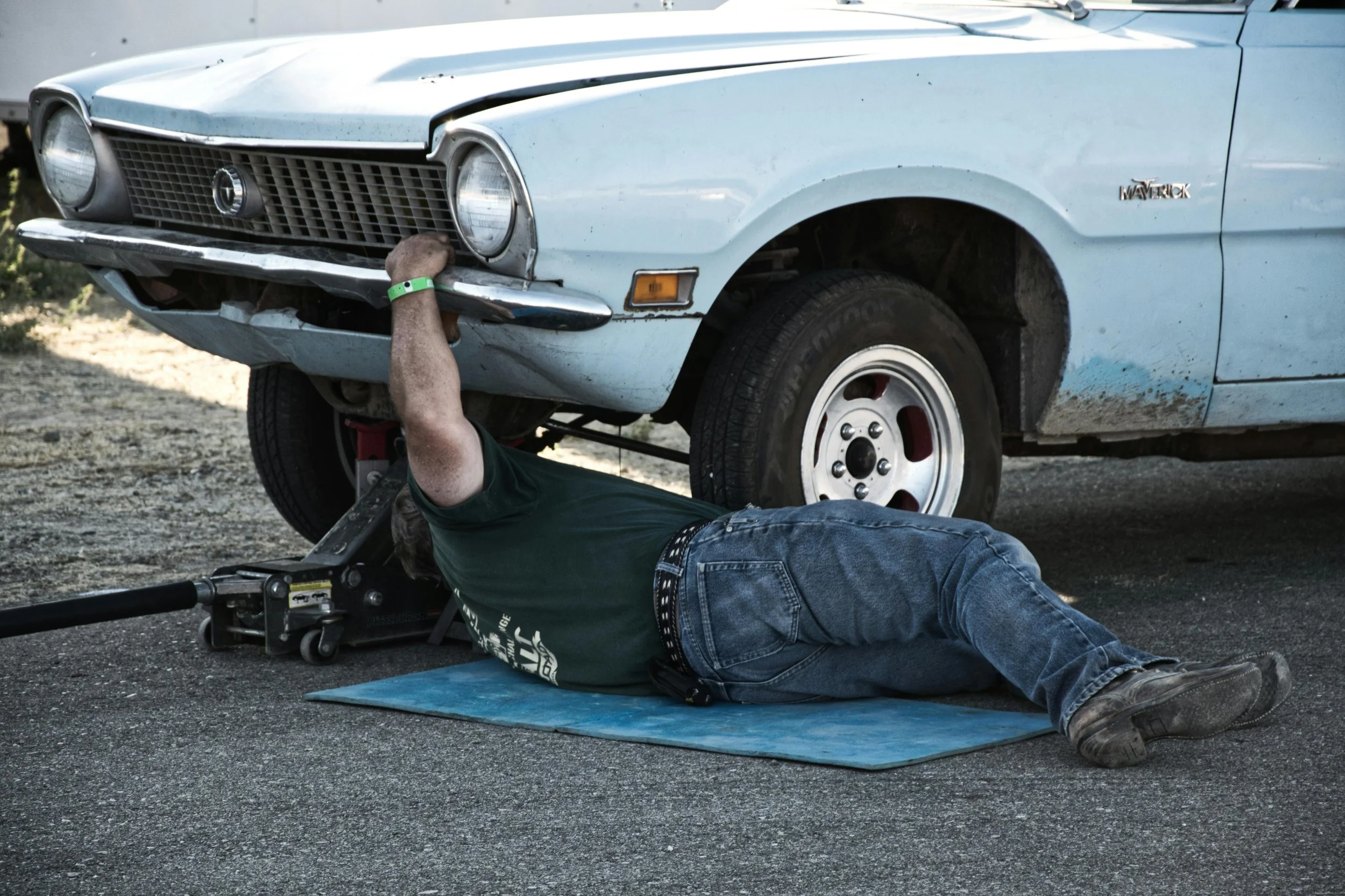 Person fixing a light blue vintage car with a wrench while lying on a blue mat on the ground.