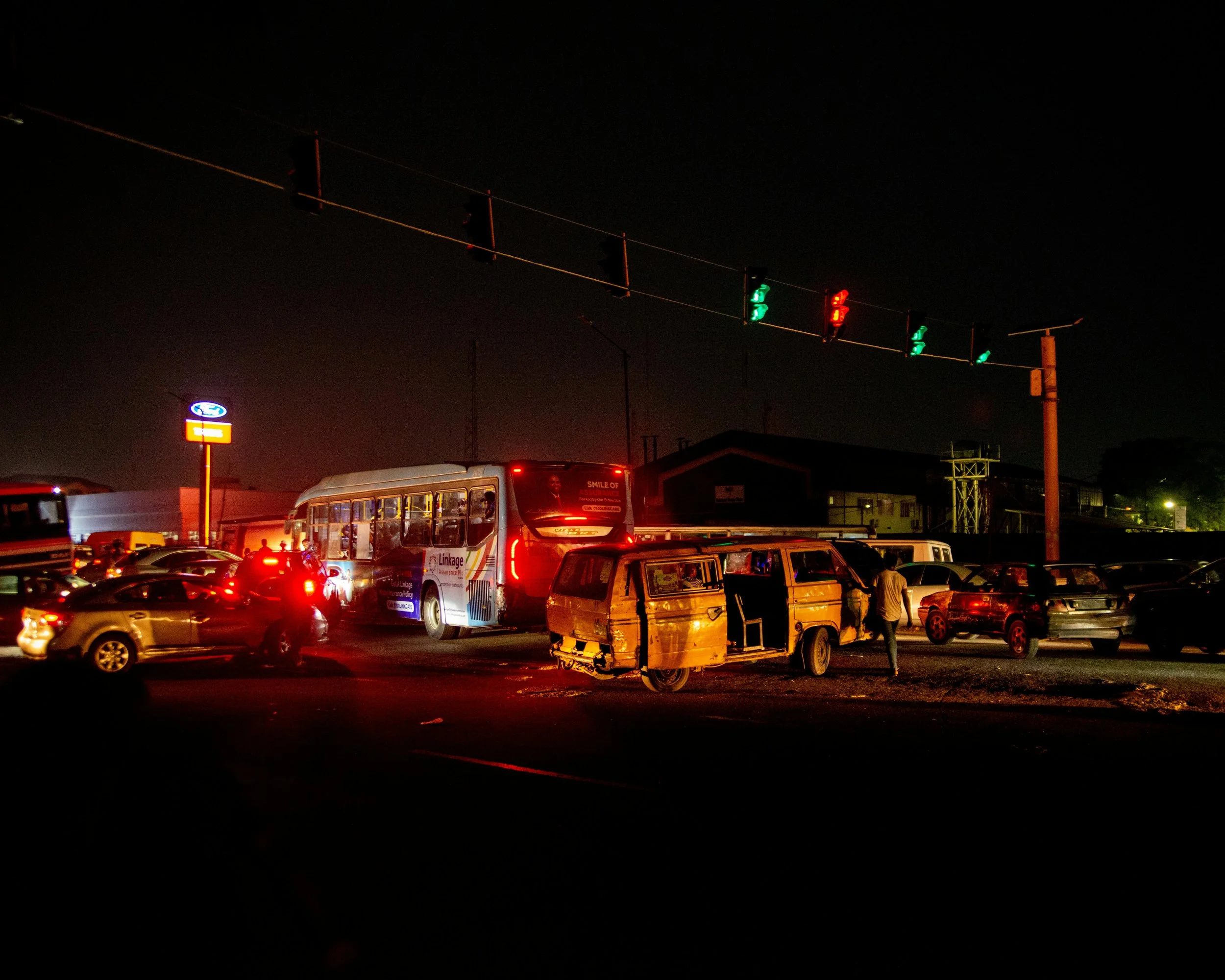 Nighttime street scene with multiple cars, a bus, and a dilapidated van under green and red traffic lights.