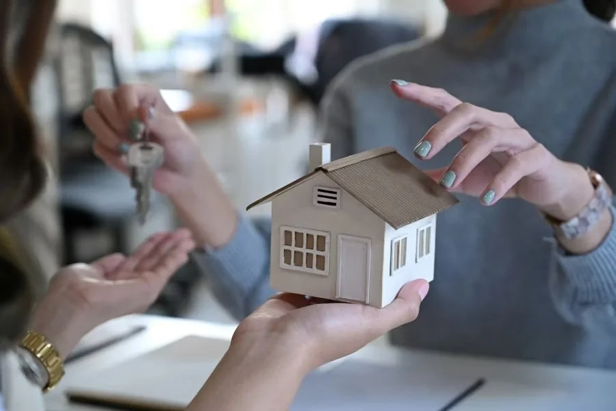Two women exchanging a house-shaped keychain in an office setting during a real estate transaction.