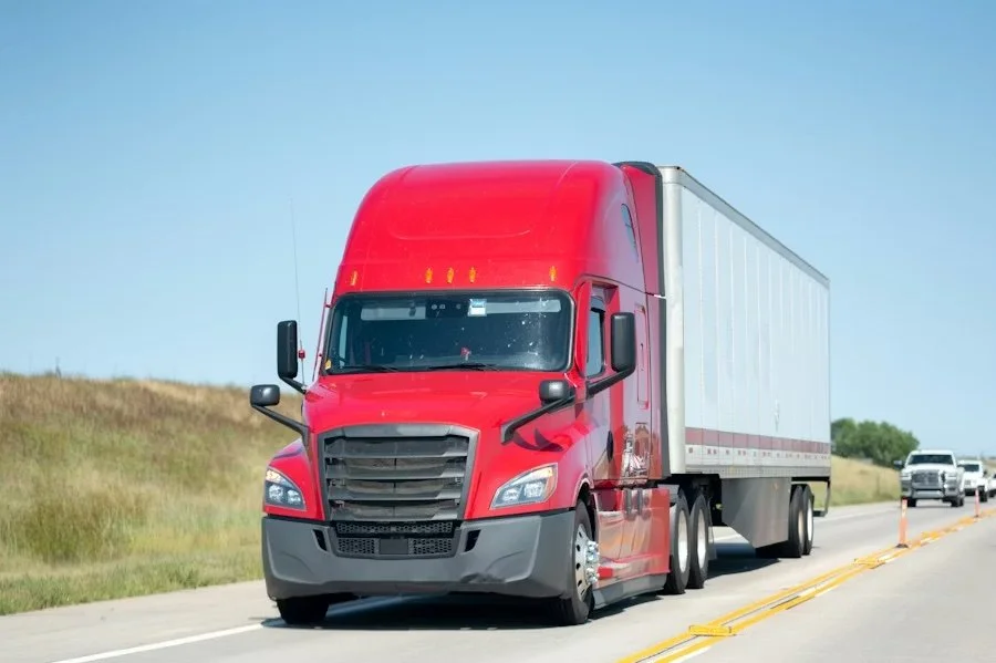 Red semi-truck driving down a highway with other vehicles in the background under a blue sky.