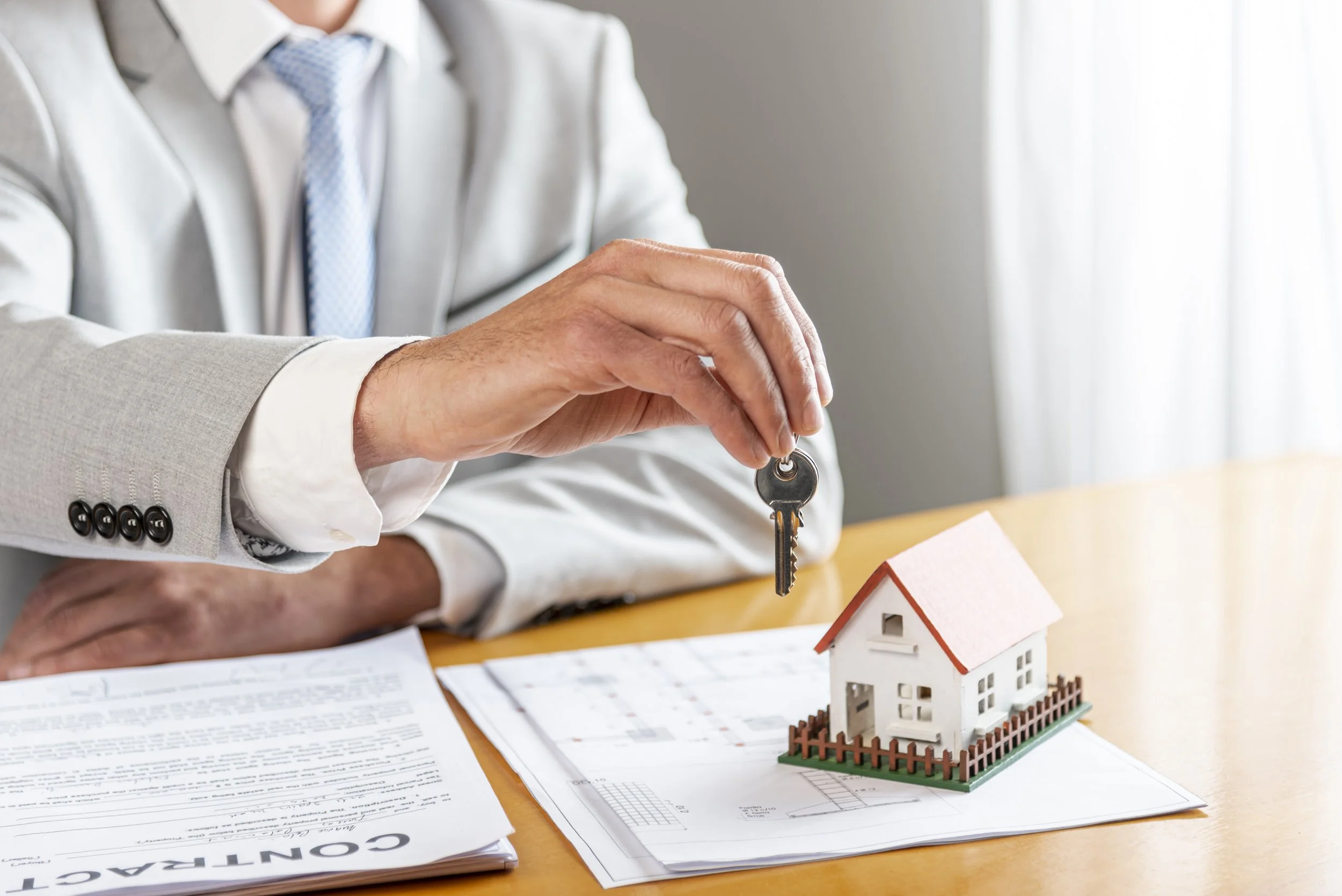 A person in a light-colored suit holding house keys above a small model house on a table with documents and plans.