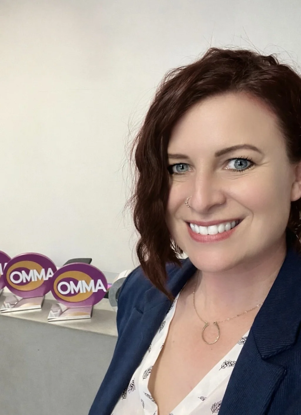 A woman with shoulder-length reddish hair, blue eyes, and a nose piercing, smiling for a selfie. She is wearing a dark blue blazer and a gold necklace. In the background, there are three purple and yellow awards with the word 'OMMA' on them placed on a table.