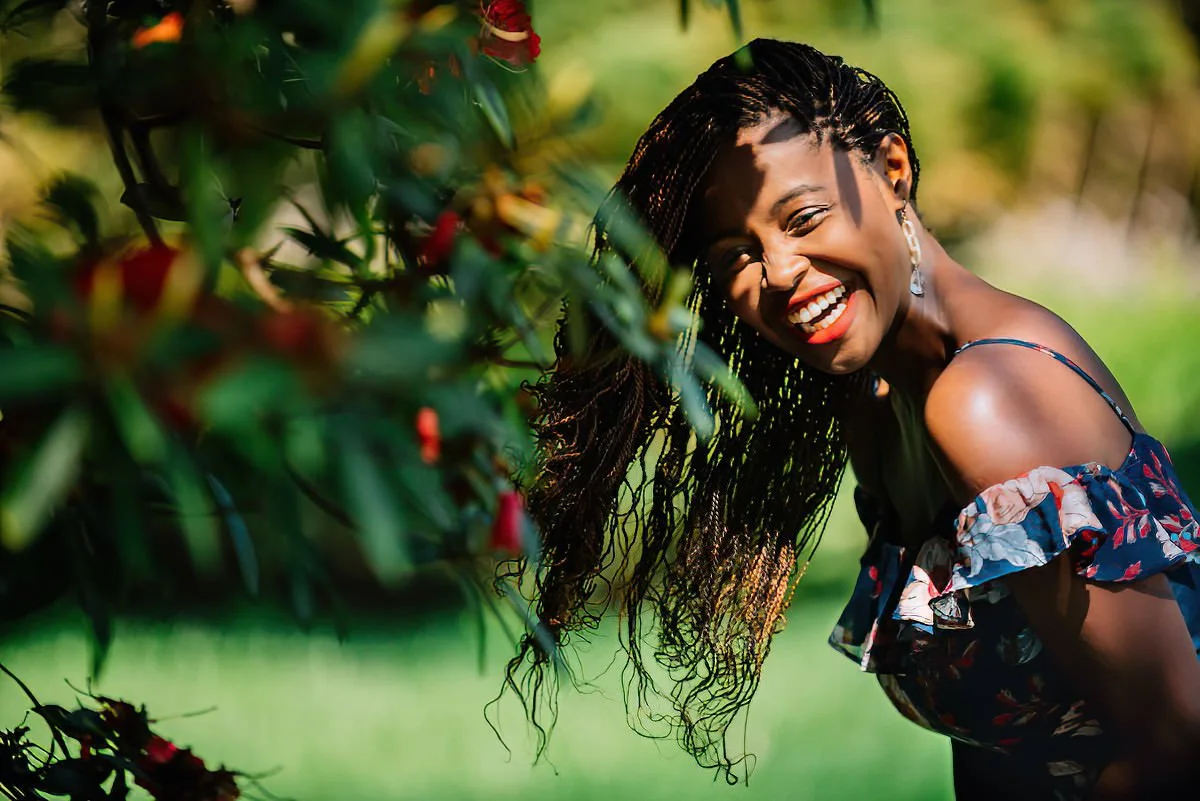A young woman with long braided hair, smiling and leaning across a vibrant outdoor garden or park area with green foliage and red flowers, wearing a floral off-shoulder dress.