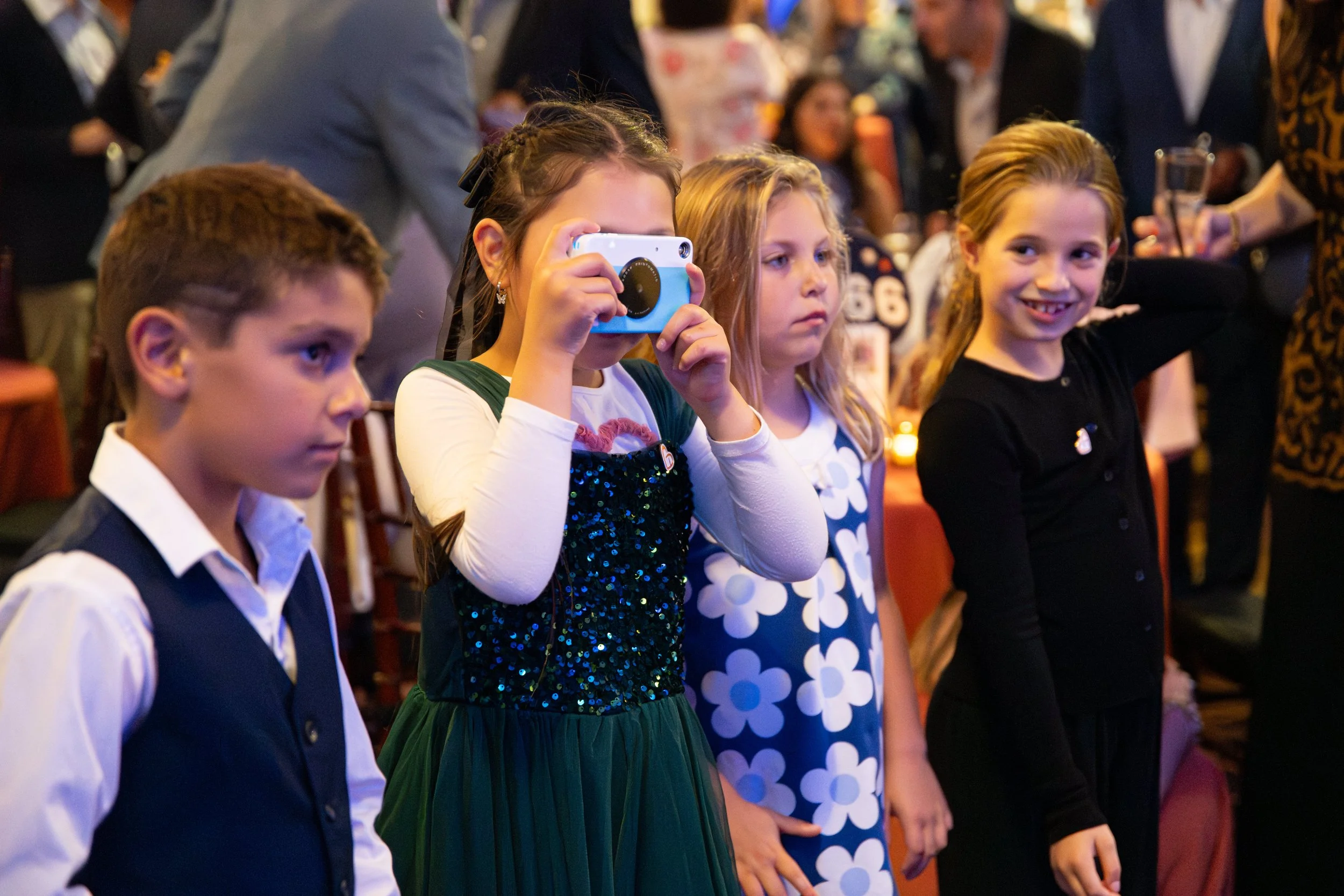 Four children standing in line, with the girl in the middle taking a photo with a camera, at a crowded indoor event.