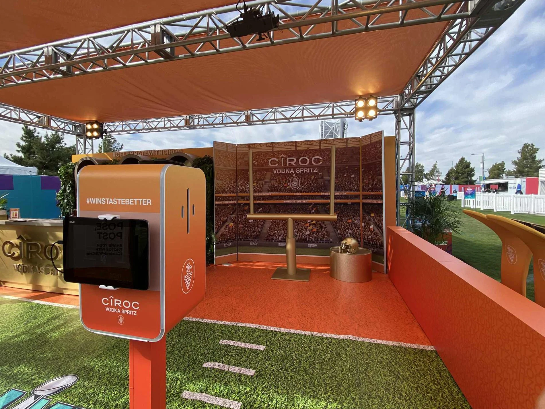 Pop-up event booth with a football theme featuring a large orange refrigerator, a backdrop with a stadium and Ciroc logo, a football helmet, and a table, set outdoors with a partial tennis court and tents in the background.