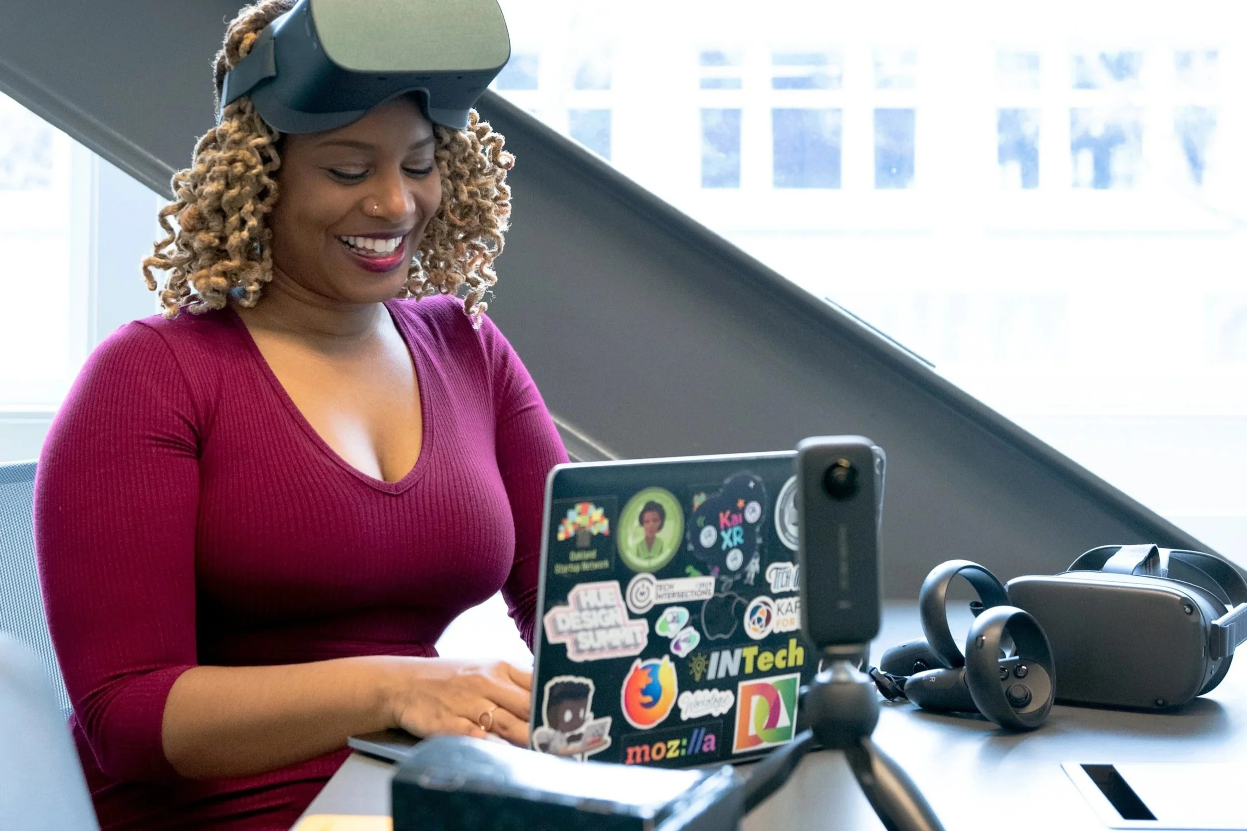 A woman with curly blonde hair wearing a red top sits at a desk, smiling while looking at her laptop, which is decorated with various stickers. She is wearing a virtual reality headset pushed up on her forehead. On the desk are VR controllers, a VR headset, and a smartphone mounted on a tripod.